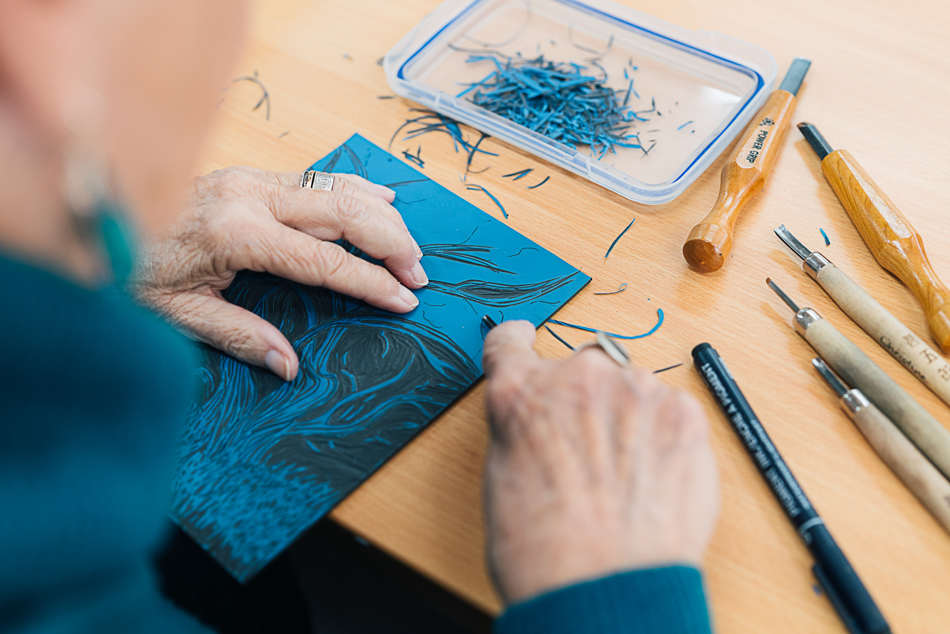 Female person wearing blue jumper carving a lino cut for printmaking using timber handled cutting tools.