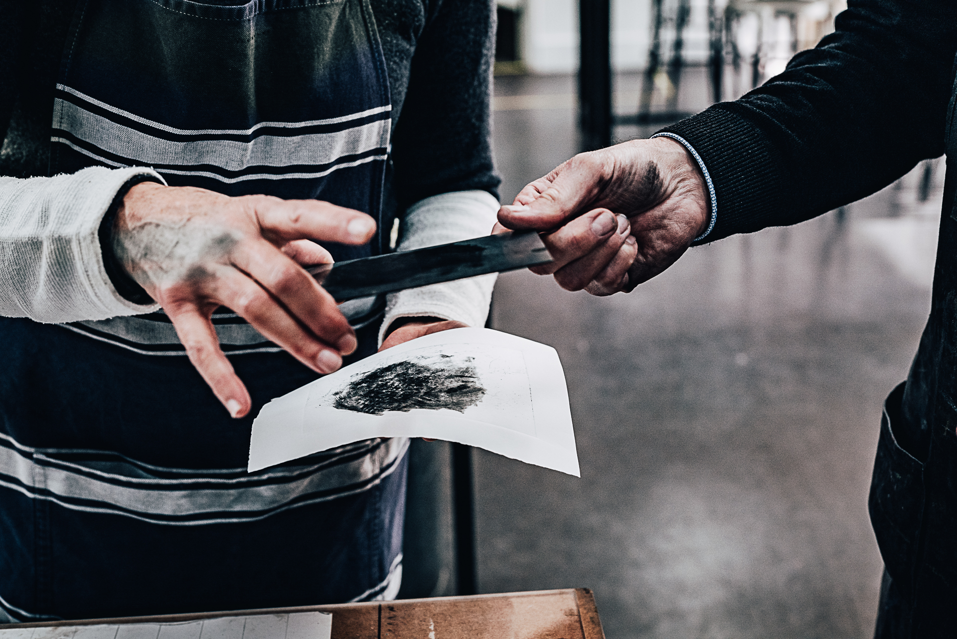 Hand of person wearing a dark jumper passing a small printing plate covered in black ink to another person who is wearing a light top and blue and white striped apron. This person is also holding a piece of white paper with a splotch of black ink in it's centre.