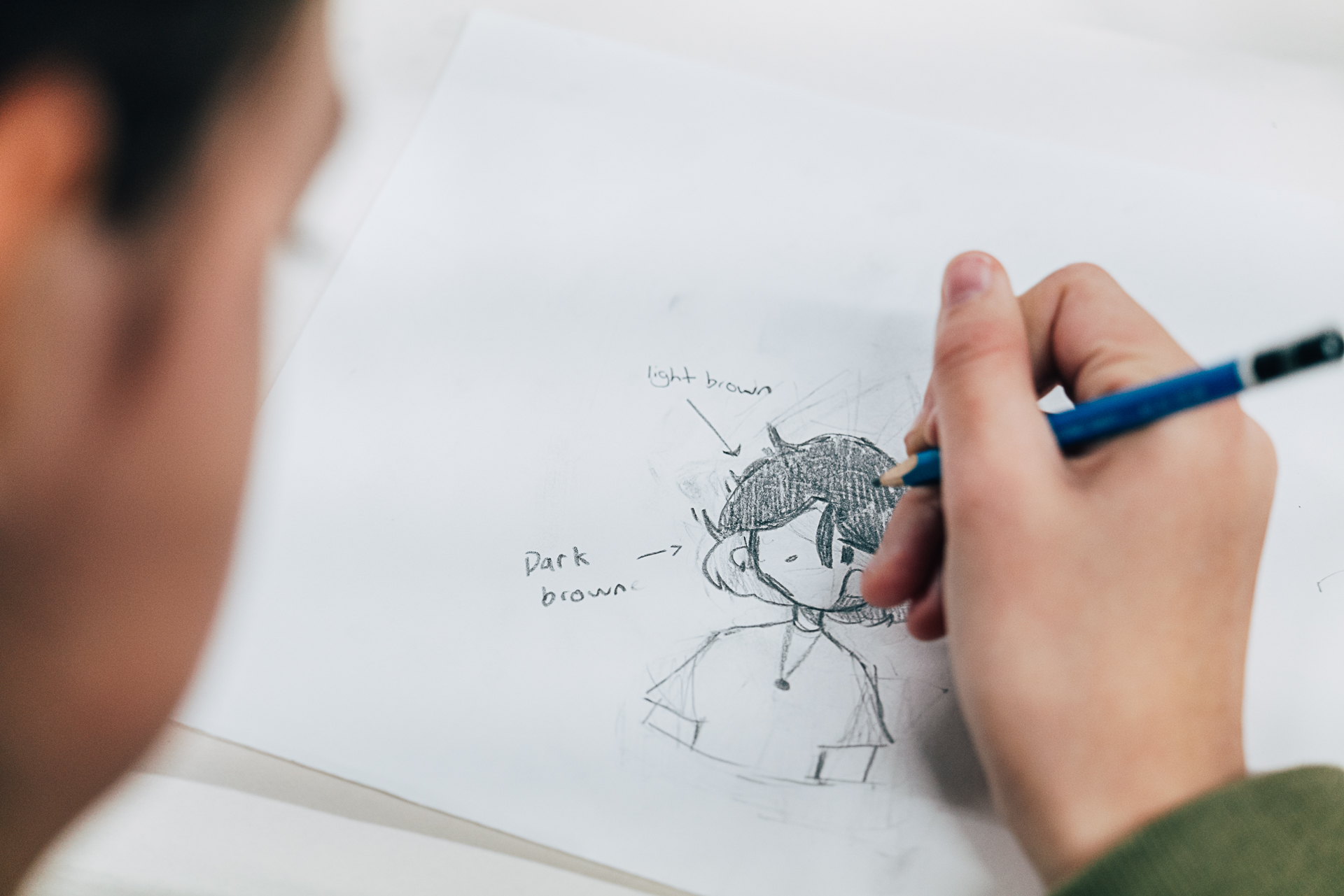 Teenager wearing green jumper drawing a character with graphite pencil on paper.