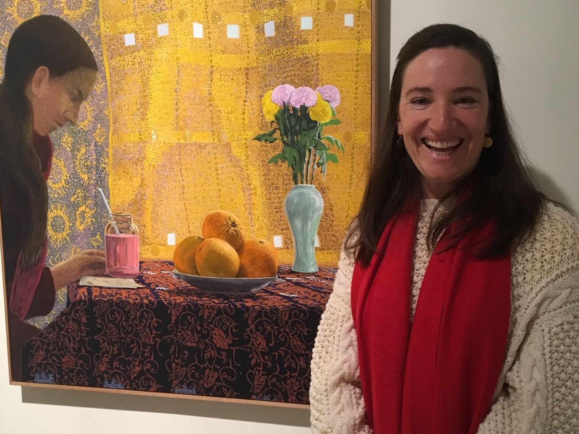 A woman with long brown hair and a big smile wearing a white knit jumper and a red scarf. She is standing infront of a painting featuring a woman sitting at a table. There is a plate of oranges and a blue vase of pink roses on the table.