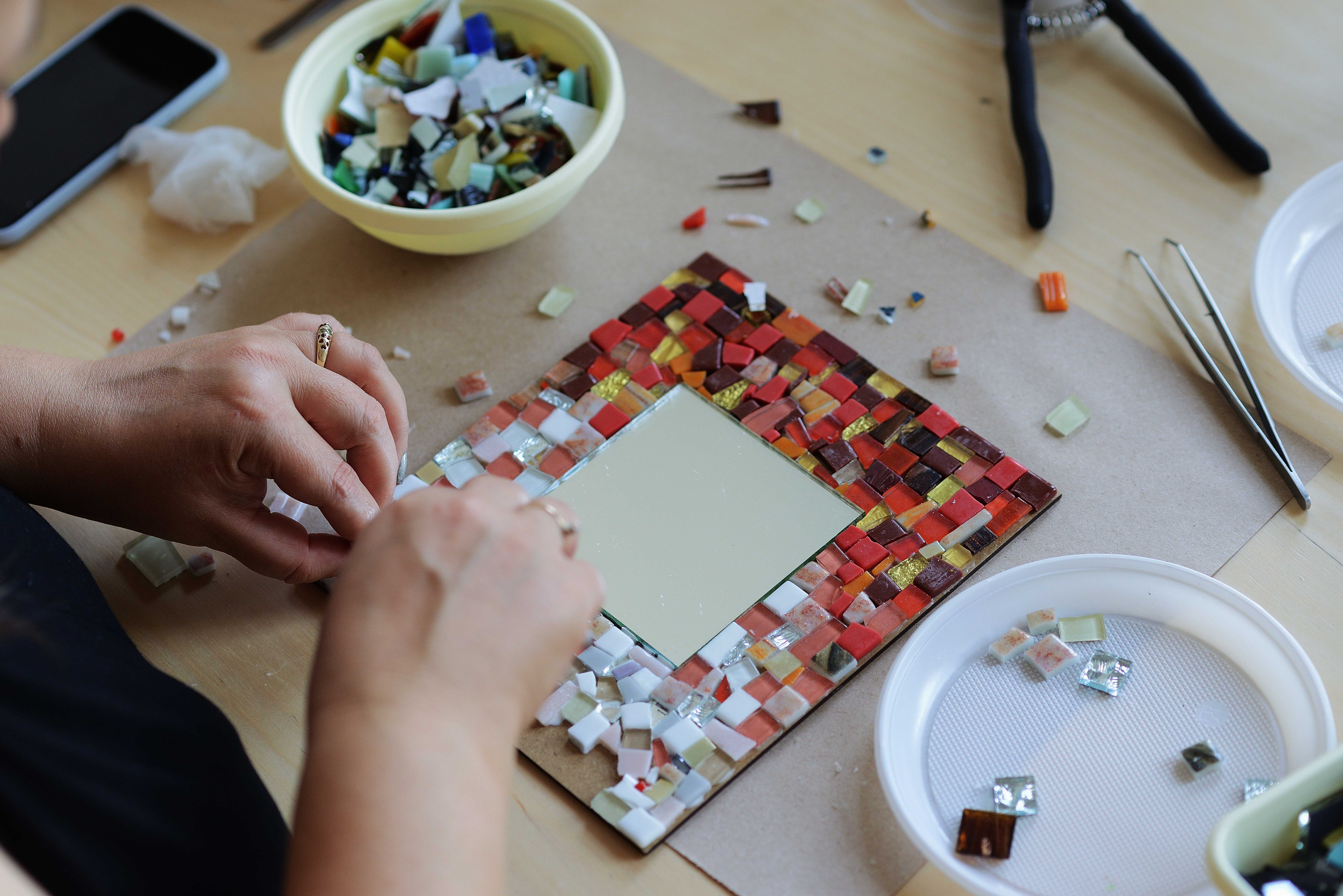Persons hands creating a small rectangular mosaic mirror frame. The mosiac tiles are small and square in shades of red, orange, yellow and white.