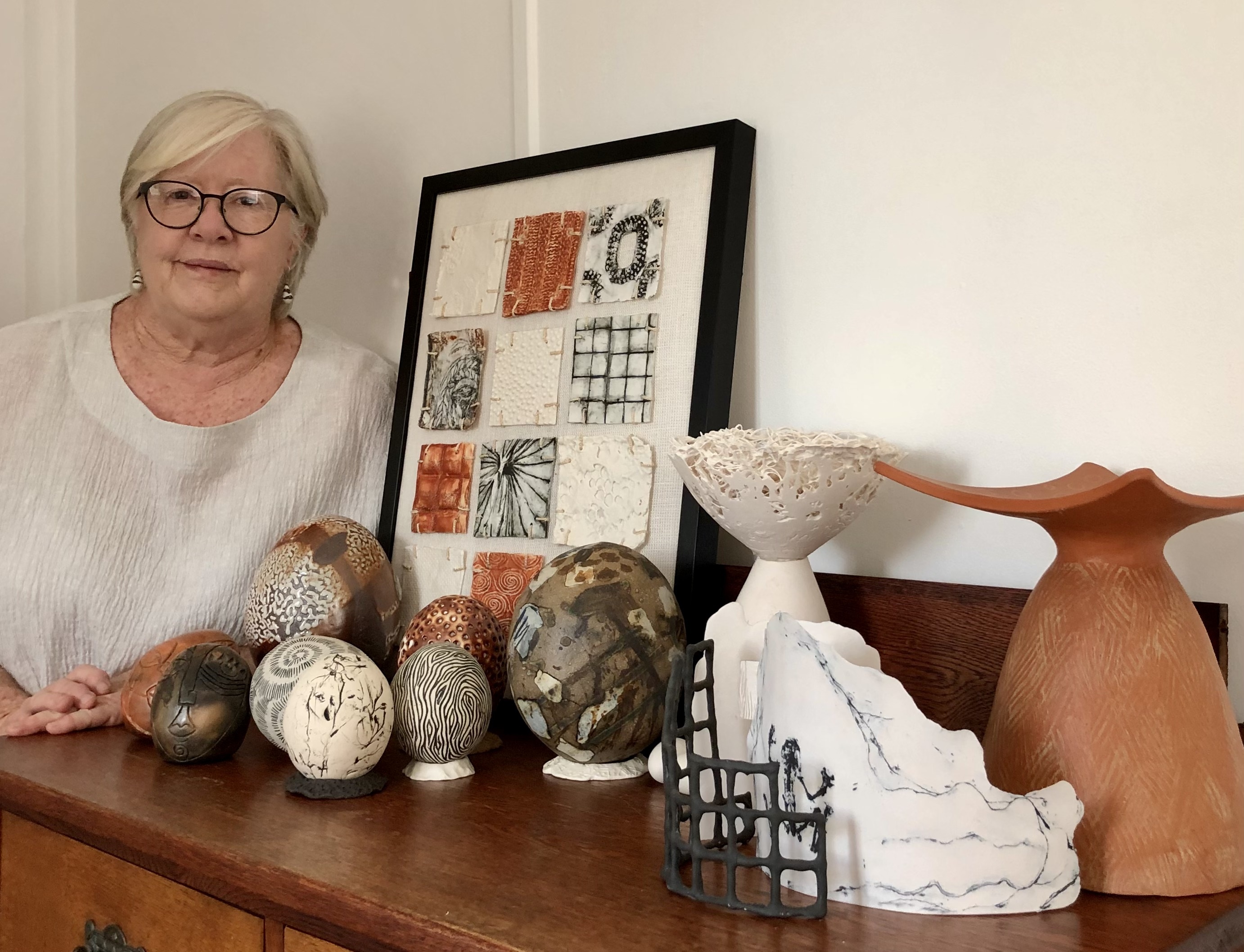 A woman with short white hair and black rimmed glasses standing behind a wooden sideboard with assorted works of pottery in earth tones on display.