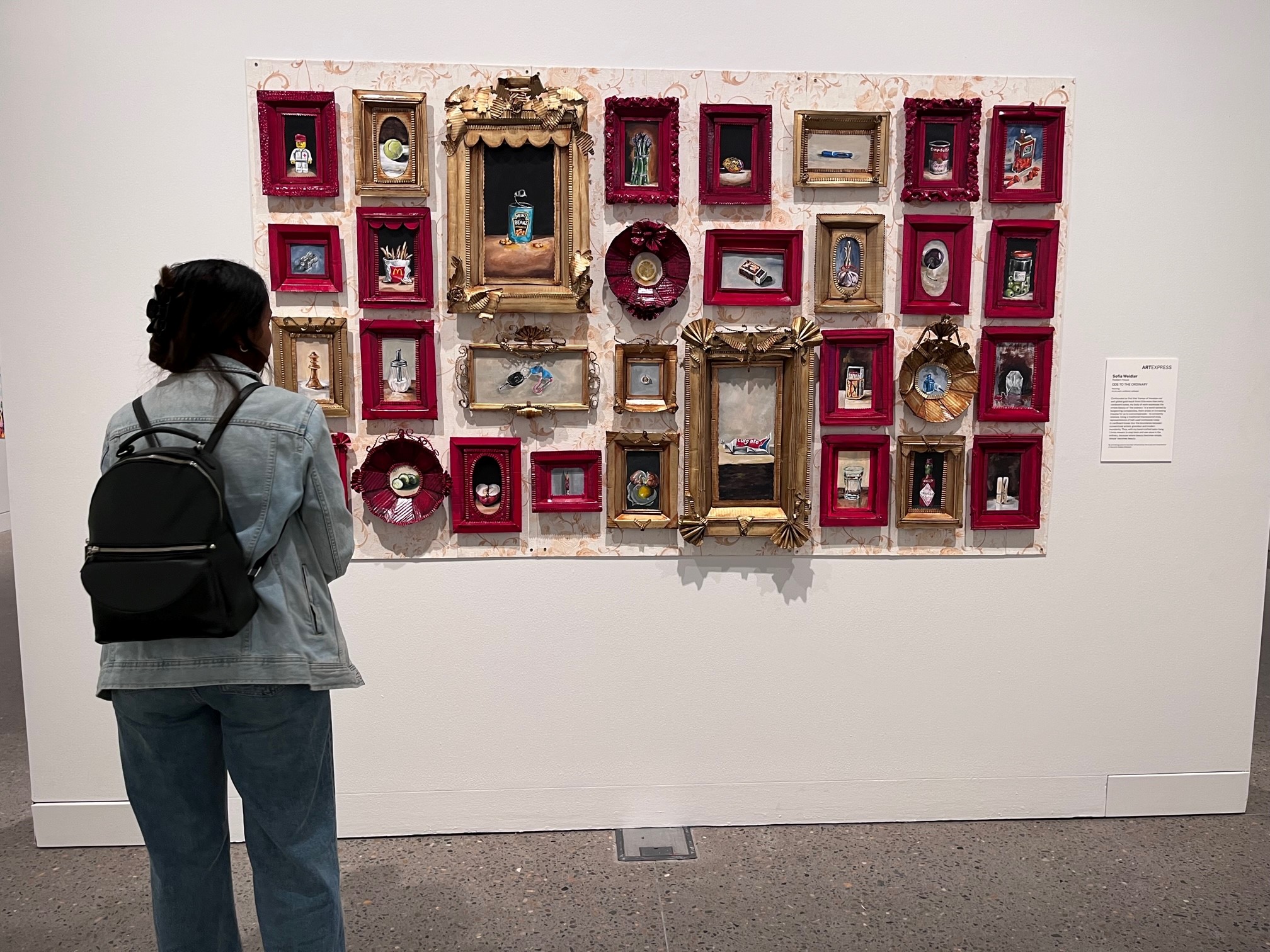 A woman with a backpack standing in a gallery looking at an artwork on the wall.