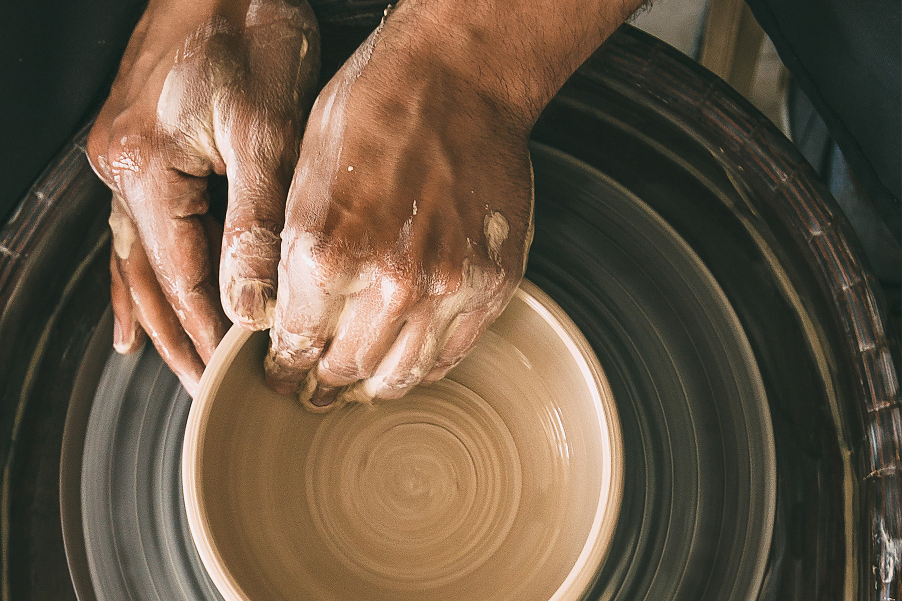 Two hands at pottery wheel throwing a bowl from brown clay.