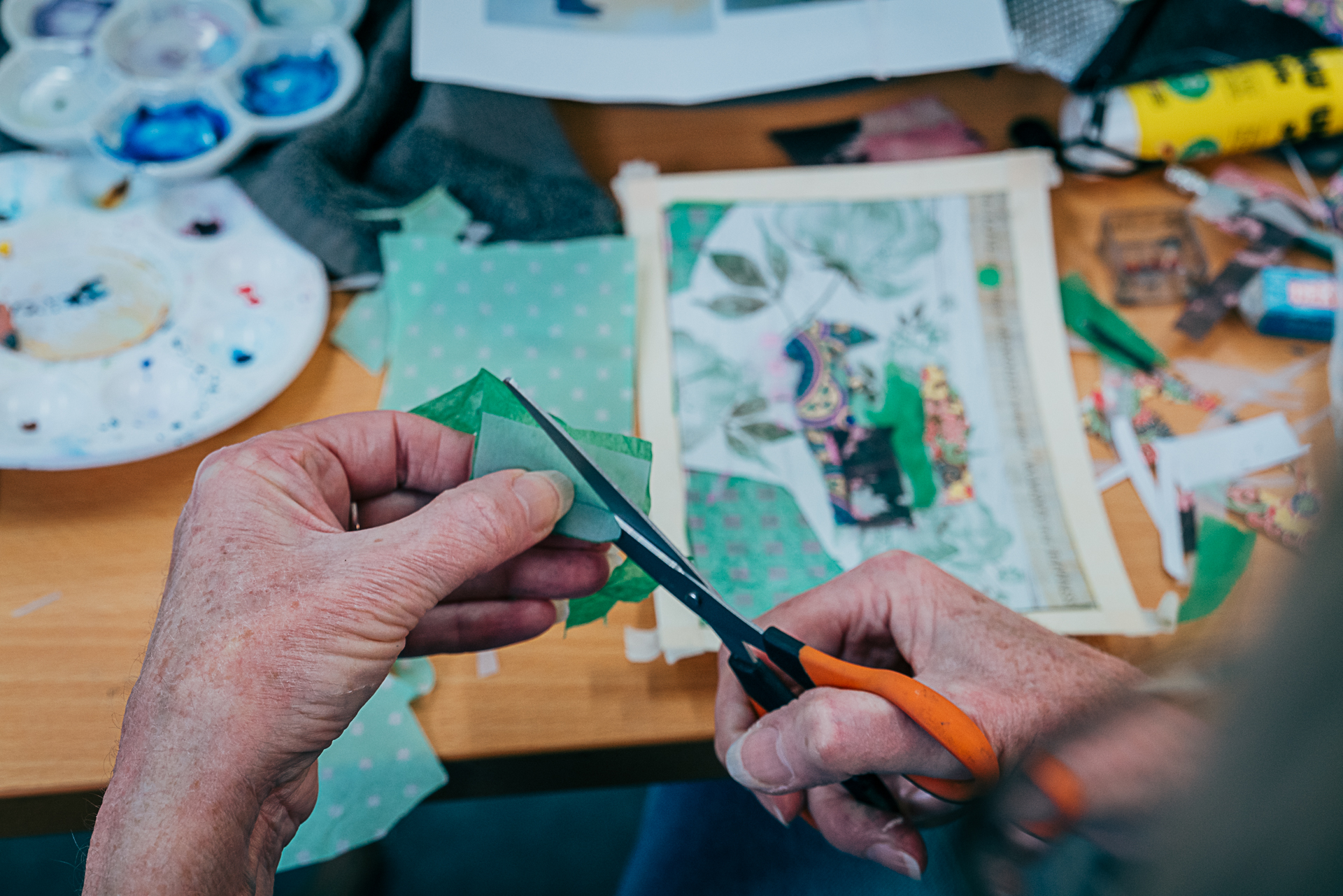 Person holding orange handled scissors cutting green paper above an in progress collage.