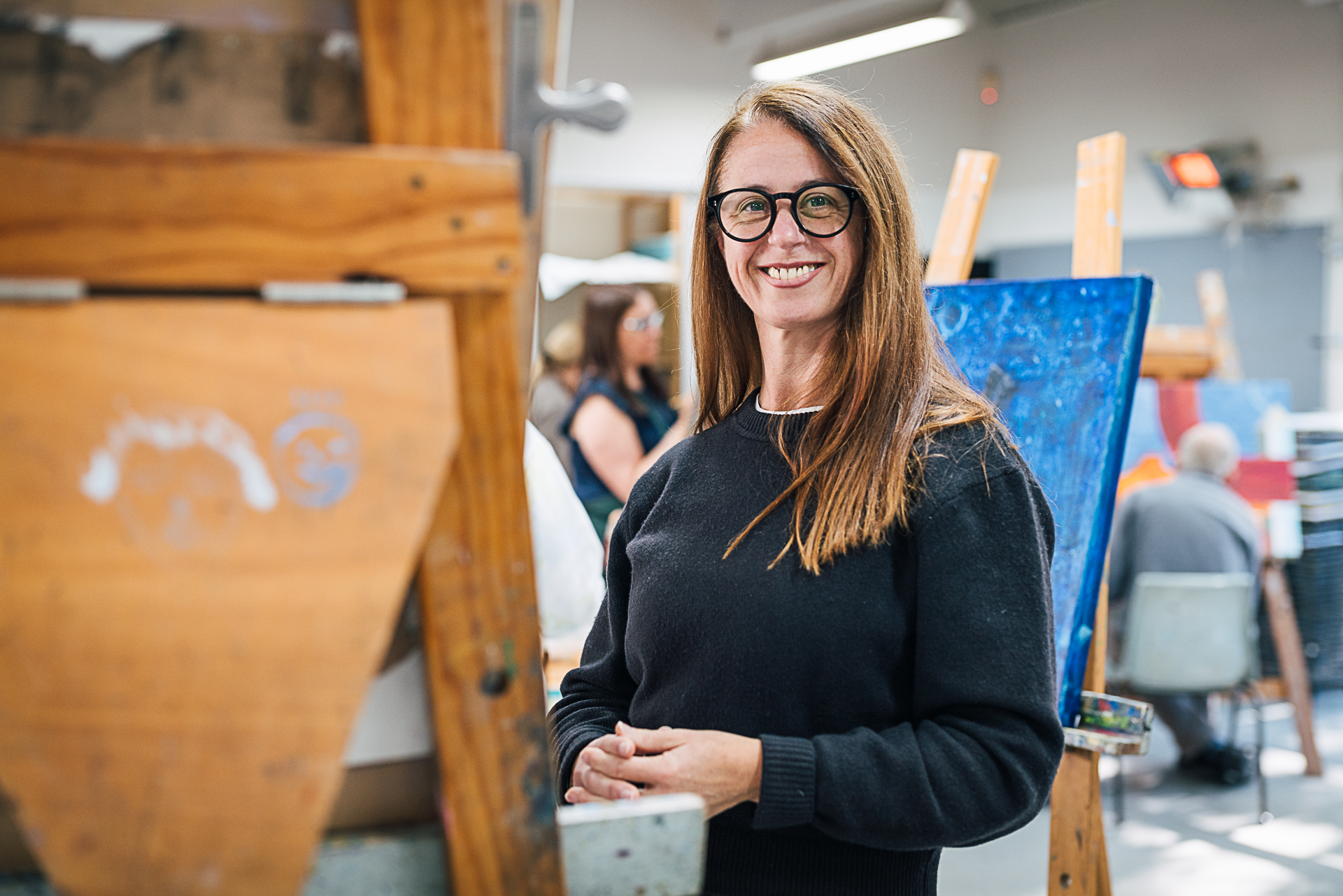 Artist and Hazelhurst teacher Michelle Cawthorn standing in front of painting easel smiling at camera. She has long chestnut coloured brown hair and is wearing a navy jumper and heavy rimmed glasses.