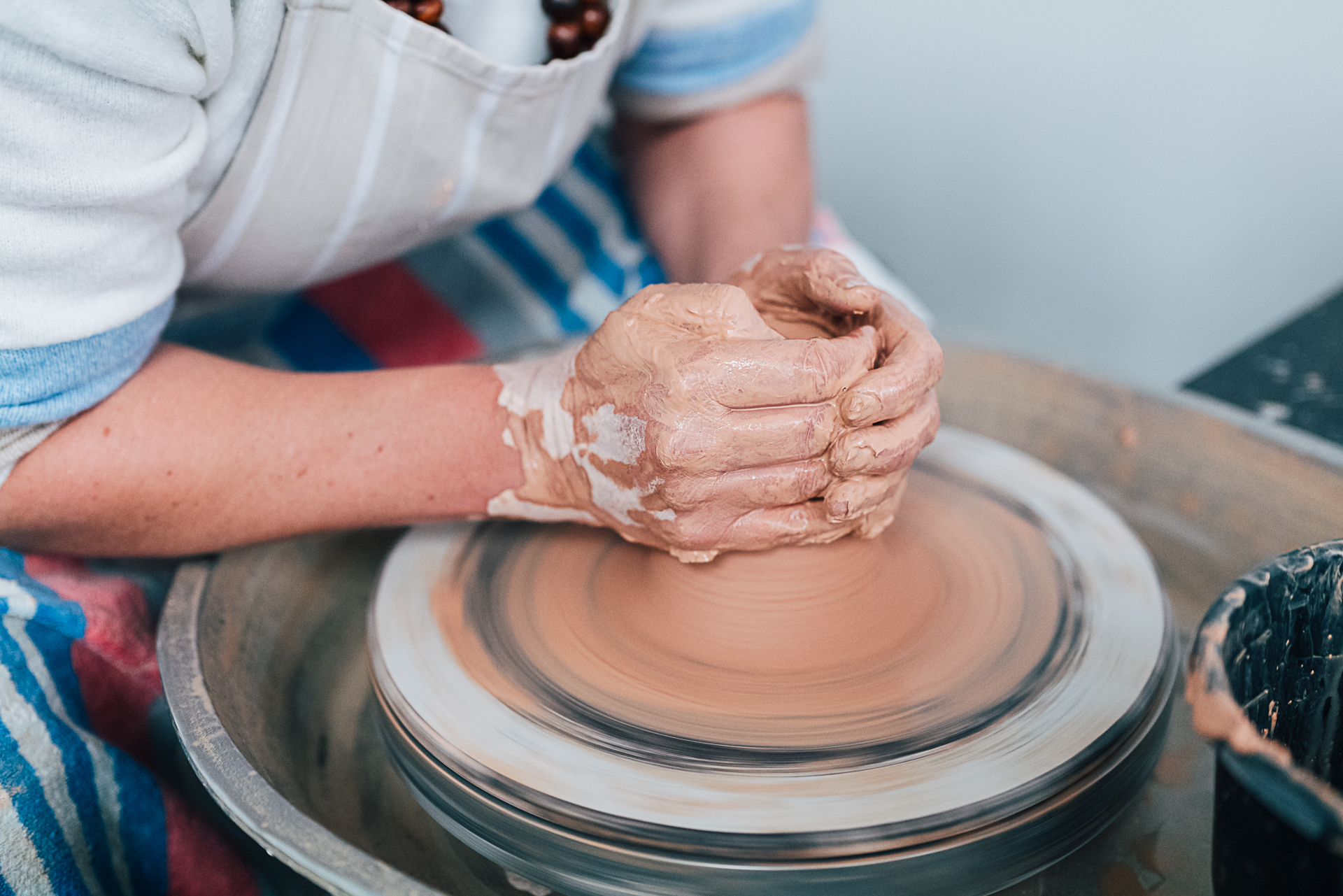 Person wearing white jumper and blue, red and white striped apron leaning over a pottery wheel, shaping red earth coloured clay.