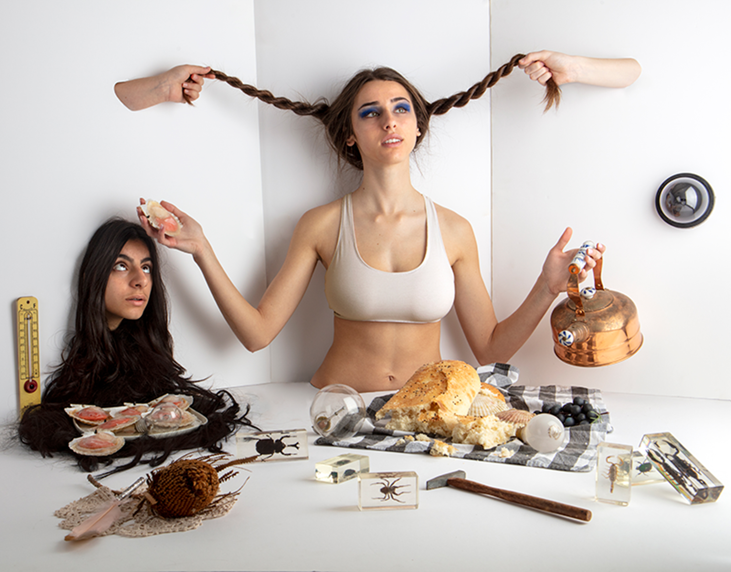 Photograph in a surrealist style, central figure is a girl with pigtails holding a copper kettle, there is another girls face poking through a wall and around them are an assortment of curious objects.