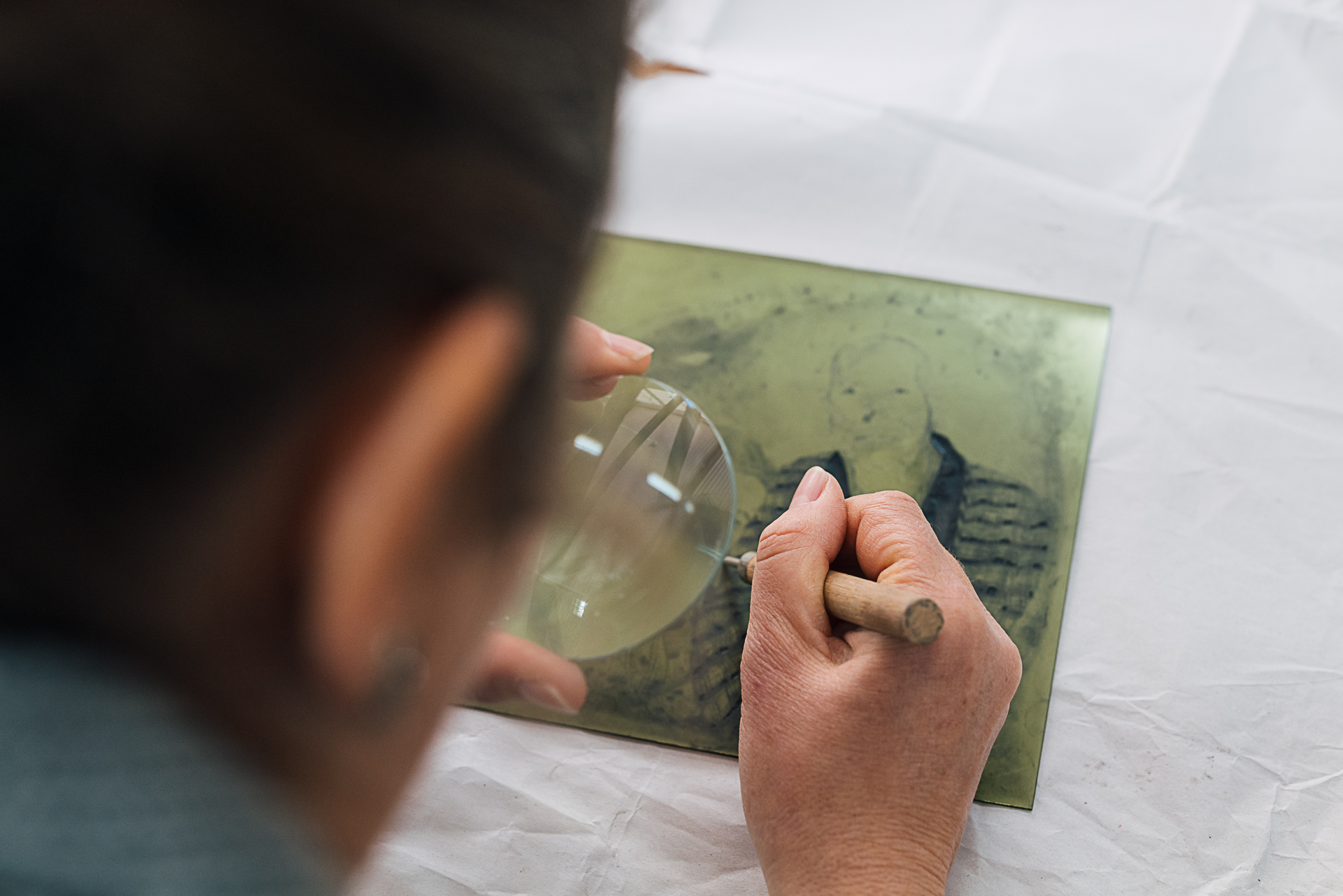 Person using hand-held magnifying glass to work on etching plate.