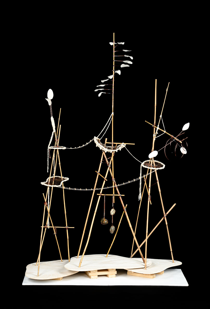 A sculpture with three peaks constructed from thin wooden sticks, the sticks are linked and adorned with other sticks and string, as well as leaves, shells and other natural elements.