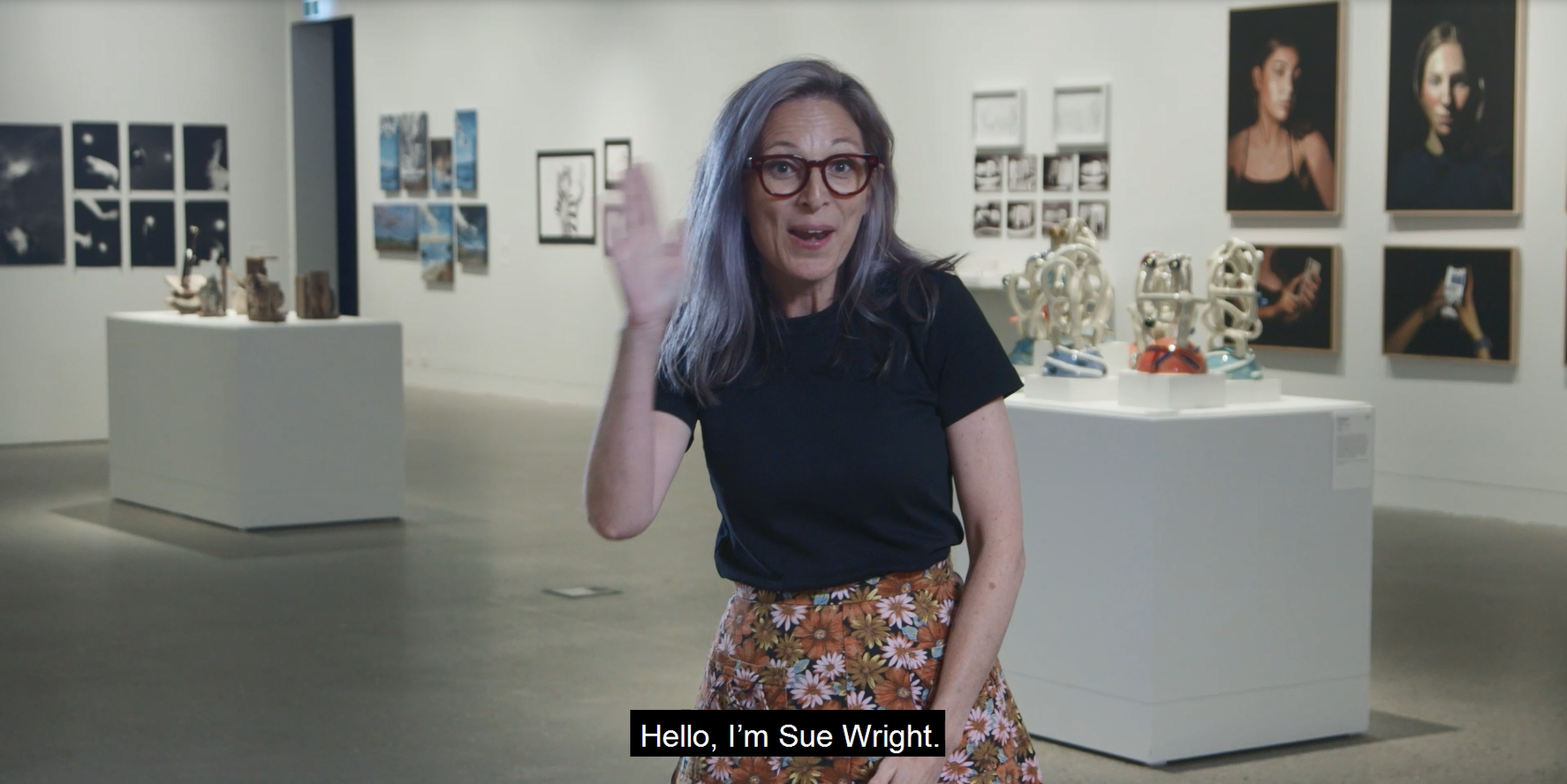 Woman with purple hair standing in art gallery. She is signing "Hello" in Auslan.