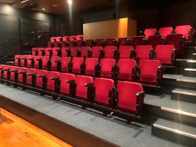 Photograph of empty theatre space with a bank of red auditorium seats, the stairs have grey carpet and the floor in front of the seats is timber. The AV desk at the top of the seating is also timber fronted.