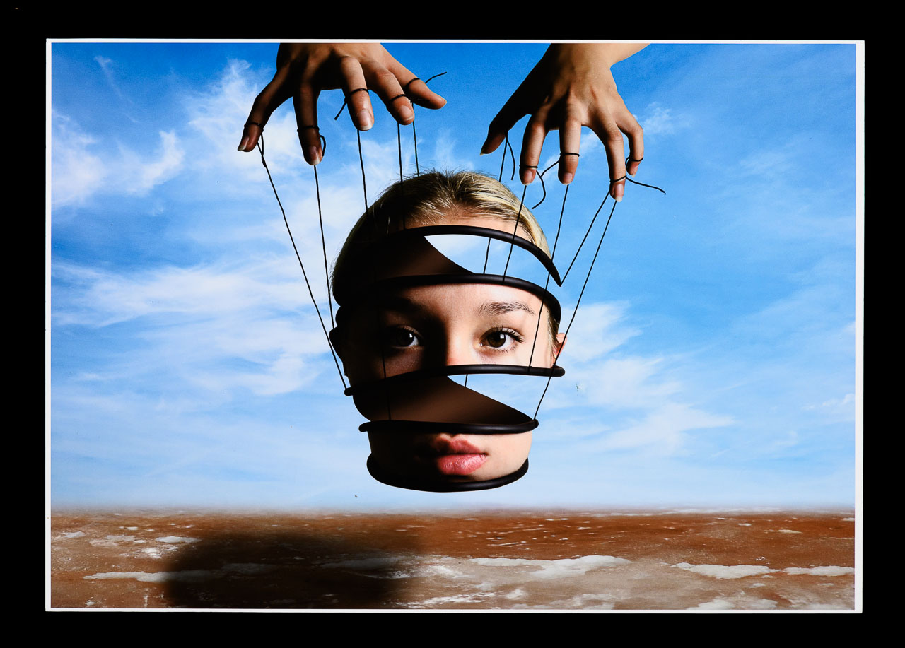 A photo media work of a female face in the shape of a spiral with marionette strings attached to it being manipulated by hands from above.