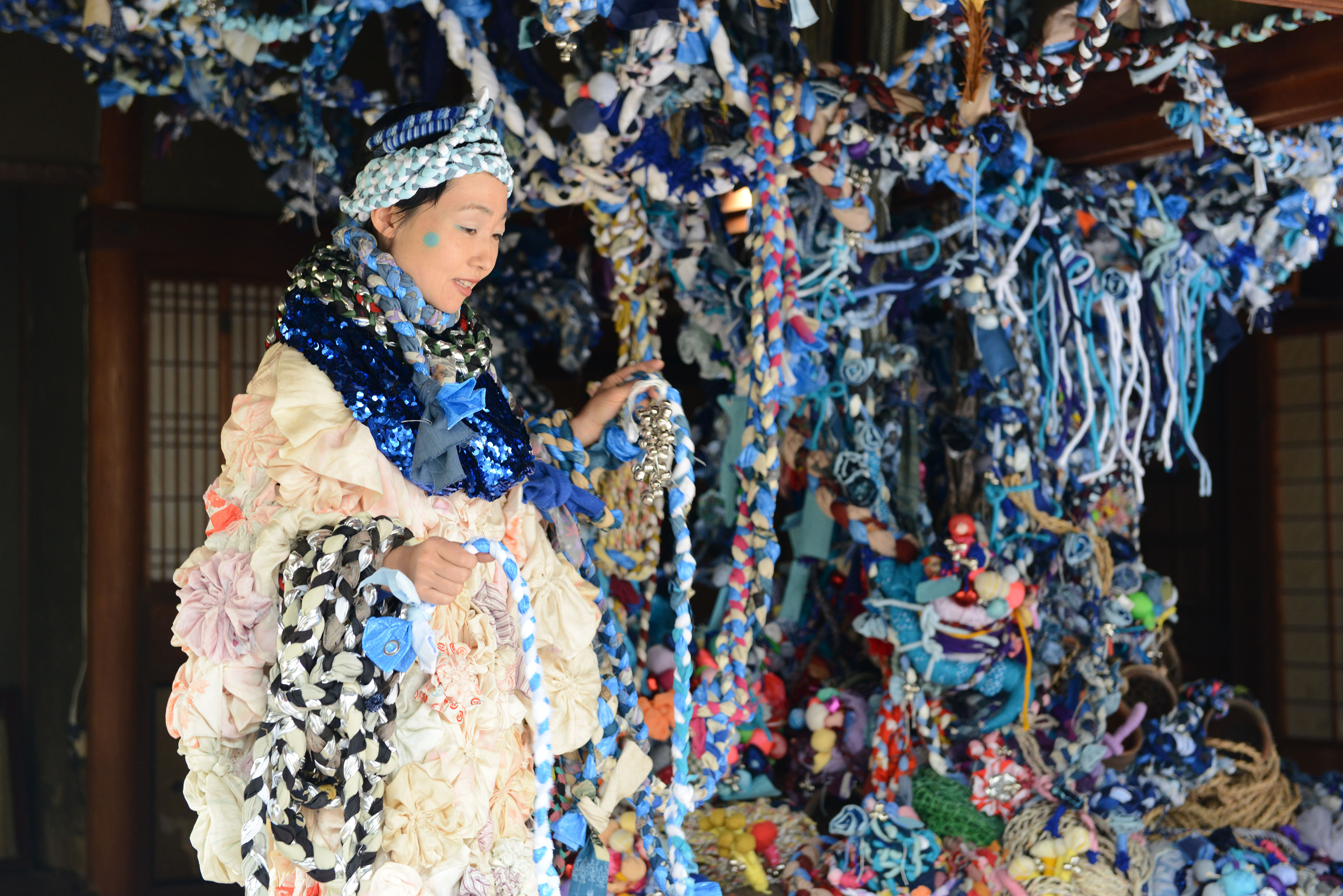 A japanese woman in an elaborate costume walking around a tree like sculpture made from blue fabrics.