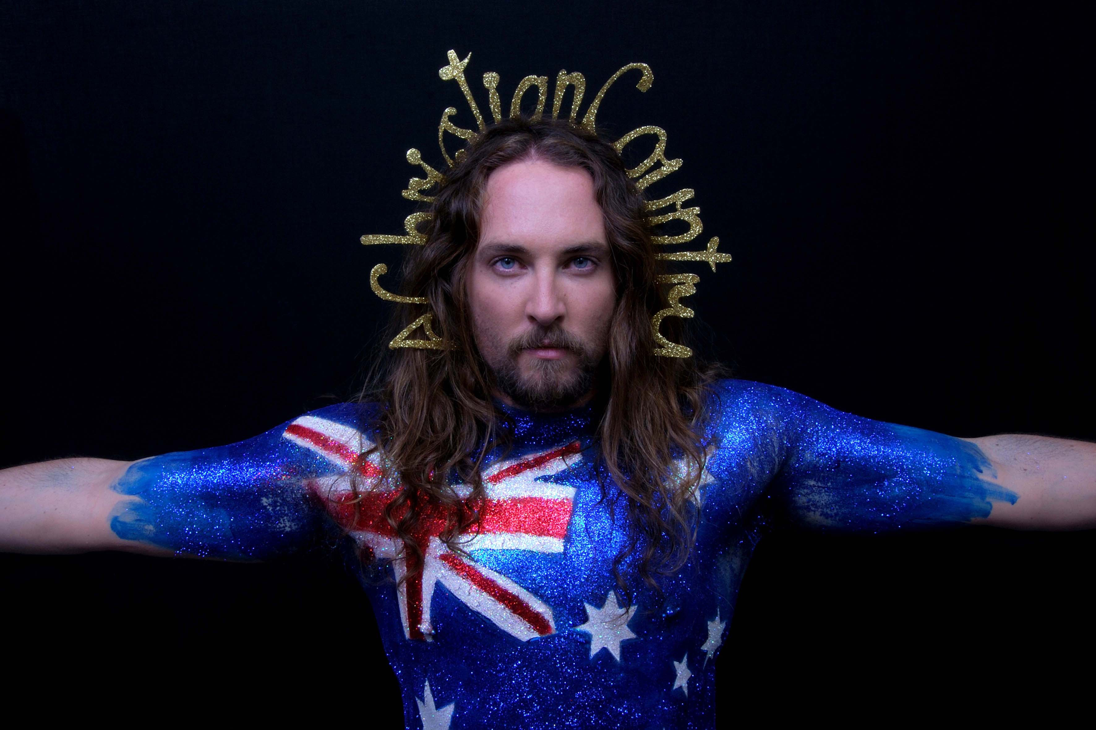 Photographic self-portrait by artist Liam Benson showing the artist's upper body with outstretched arms covered in glitter in the design of the Australian flag, wearing a gold crown of looping letters that spell "A Christian Country".