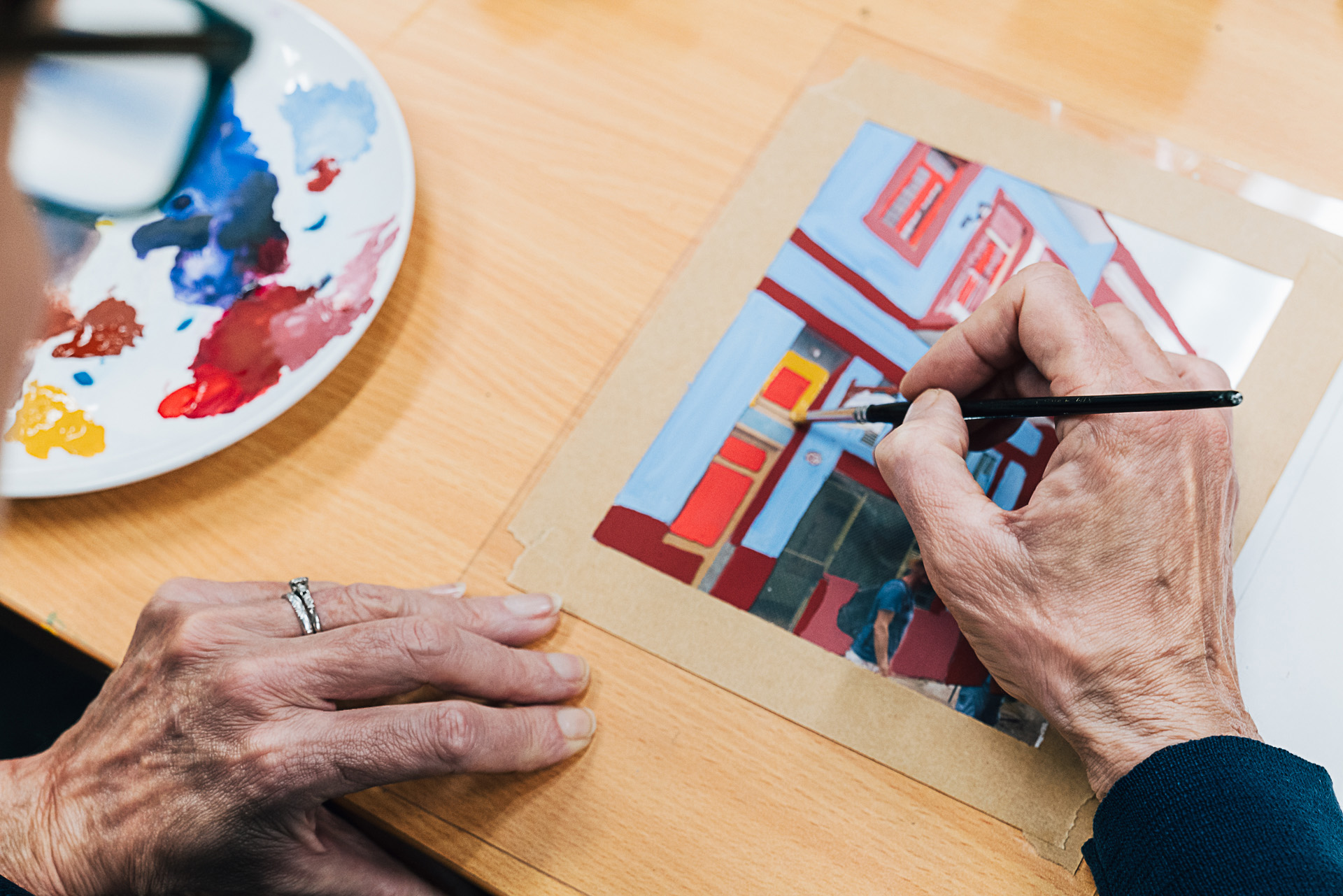 Close-up of hands painting a colourful street scene with a small brush, a paint palette visible nearby.