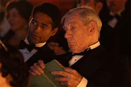 Two men in tuxedos sitting in an audience, one is white and one is black.