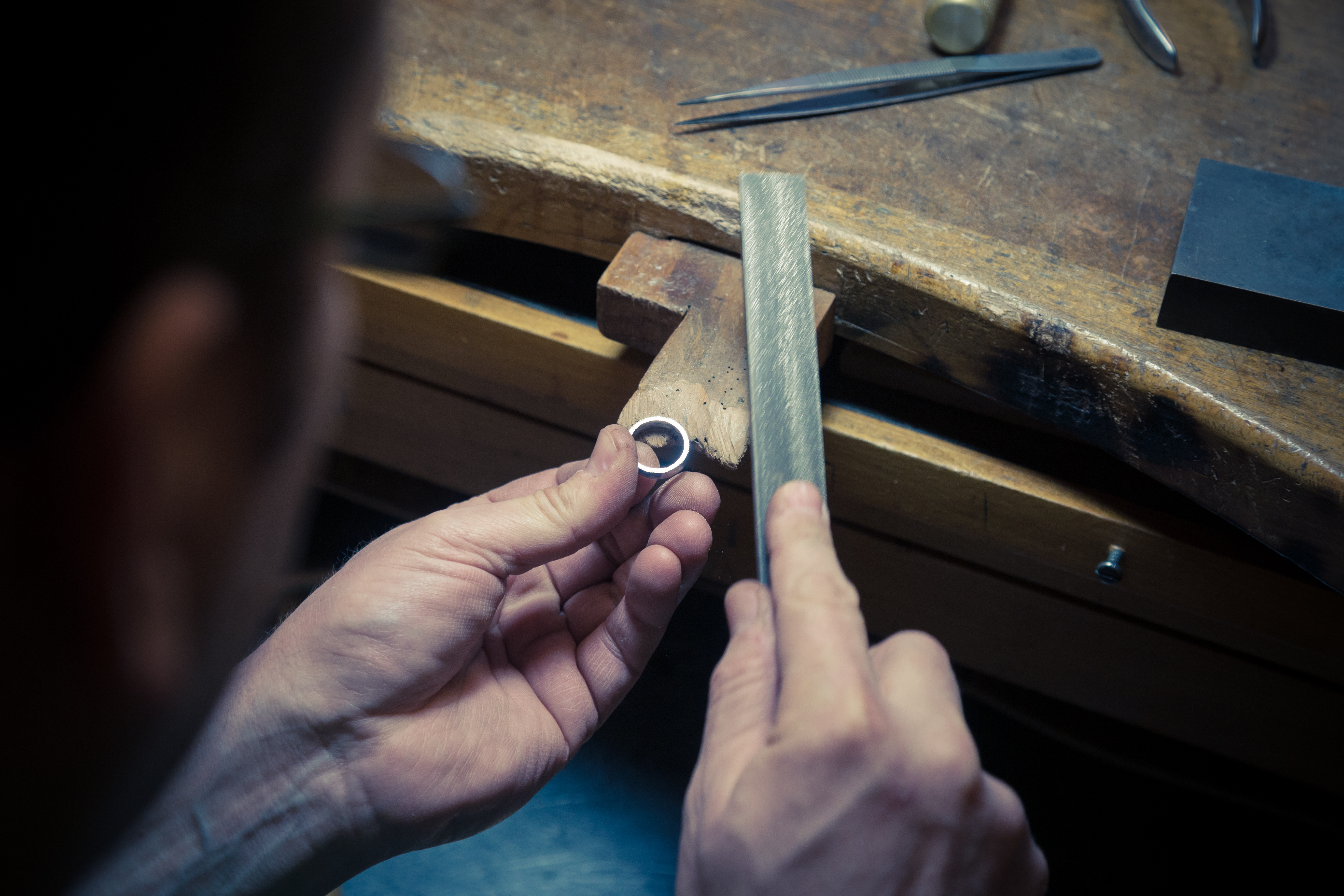Photograph looking over woman's shoulder. She is sitting at a timber jeweller's bench, filing a silver ring with a hand-held metal file.
