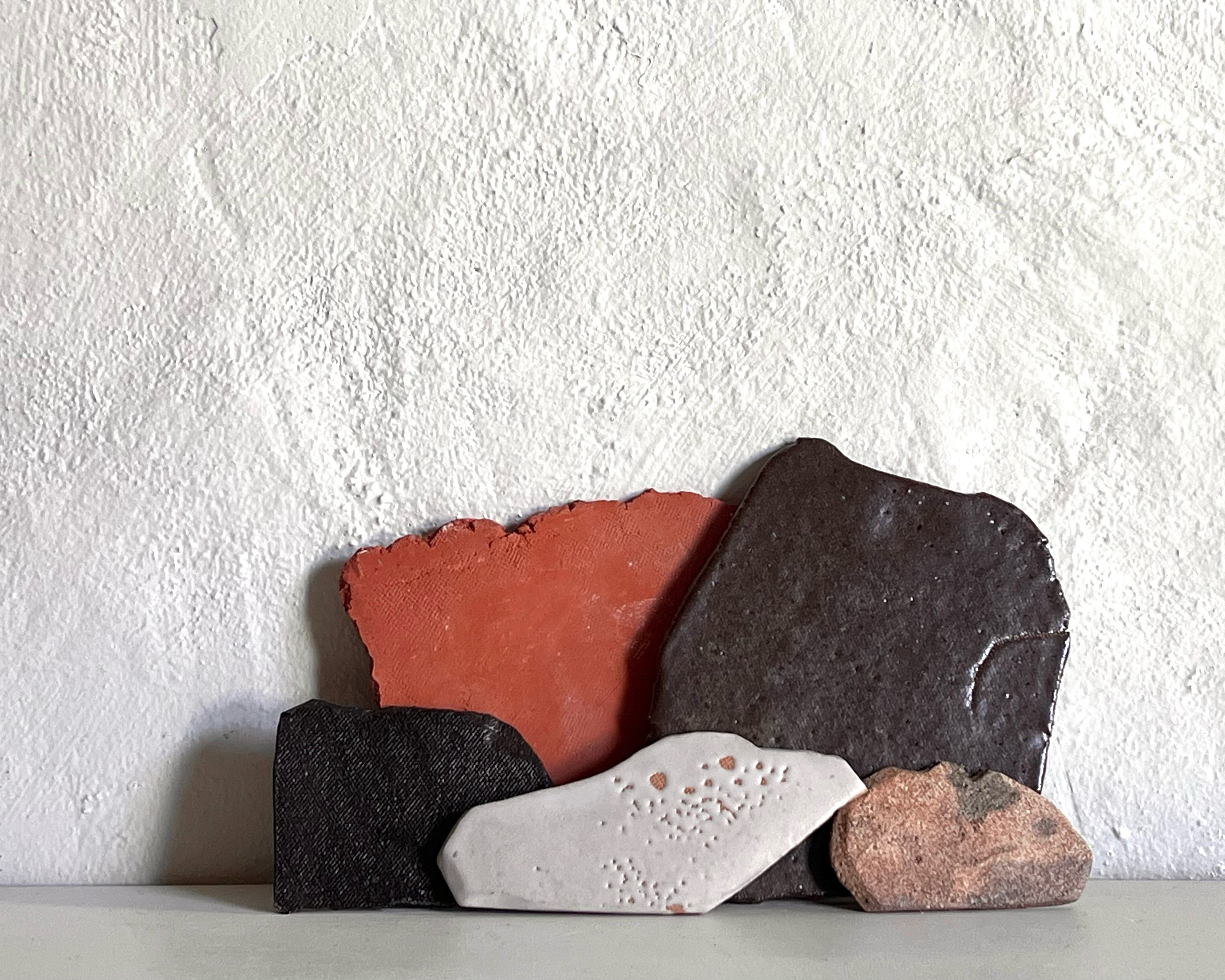 Five small organic, flat shapes of clay in earth tones arranged to form a landscape style arrangement leaning up against a wall.