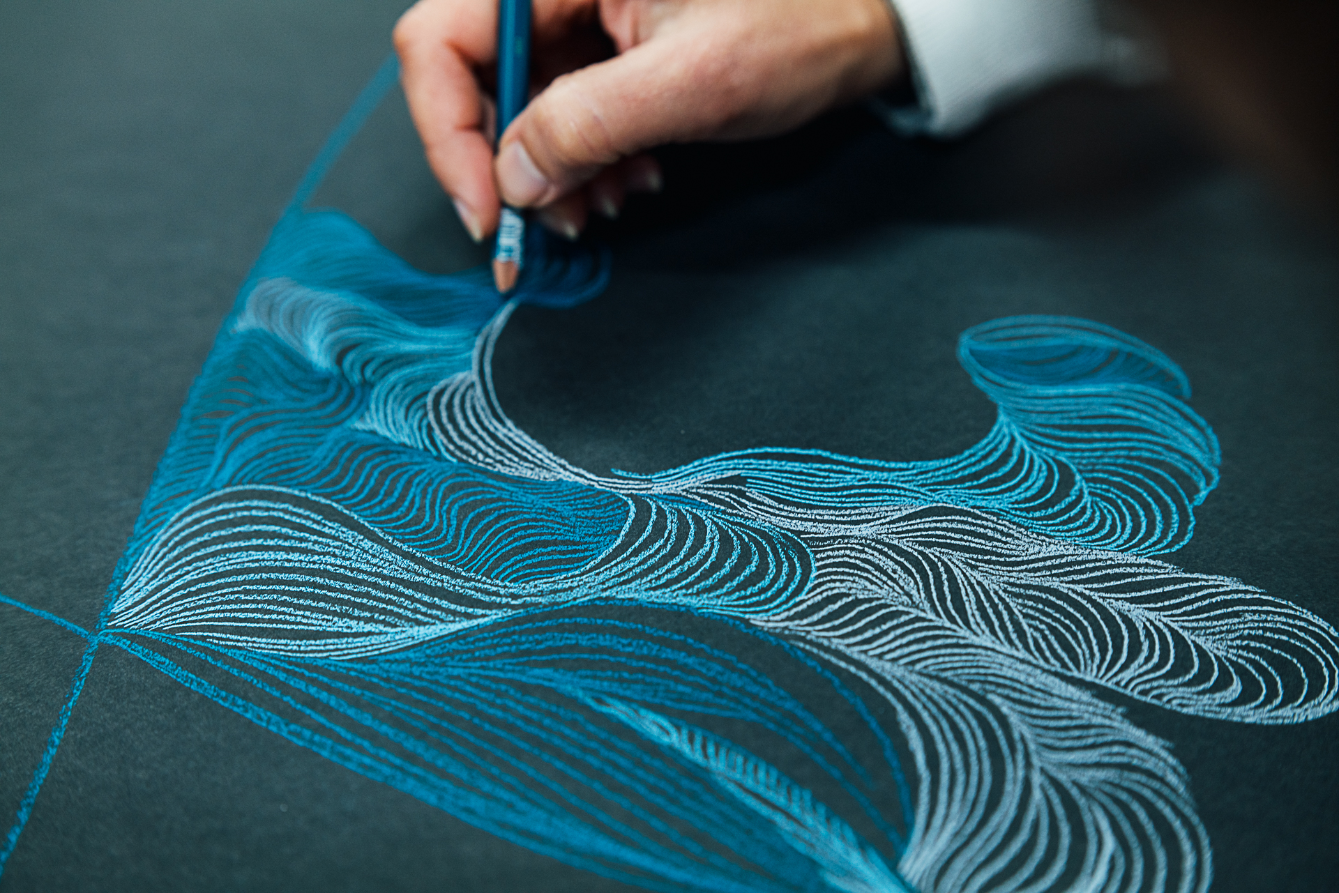 Hand holding pencils, drawing on black paper with blue pencil. Drawing features lots of wave-like, swirling lines.