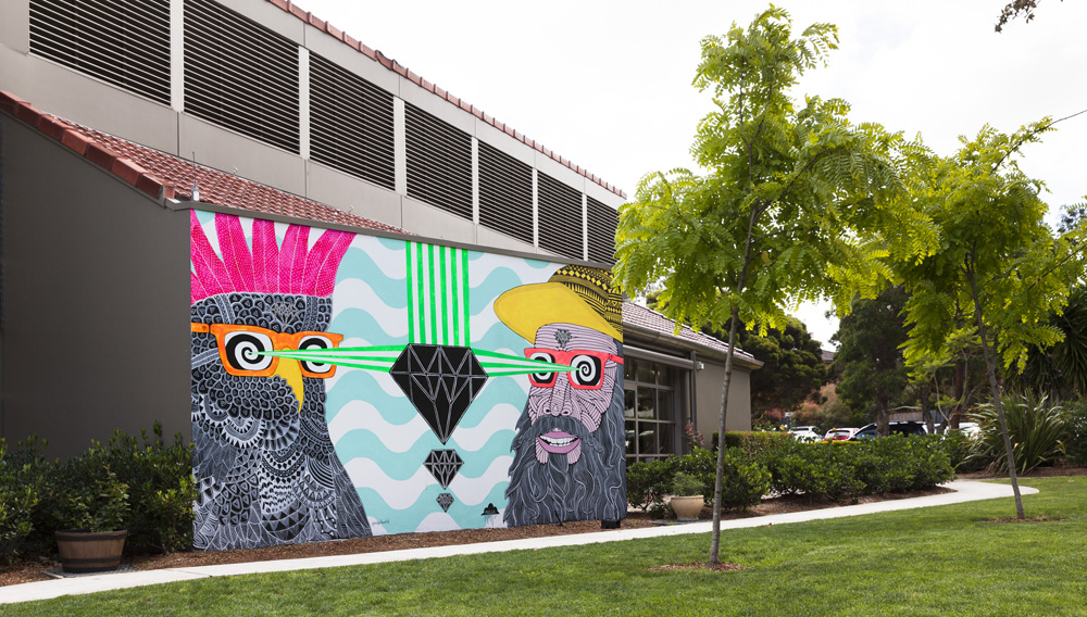 An brightly coloured outdoor mural featuring two cartoonish characters wearing orange framed glasses facing each other. One character is a bearded man wearing a yellow cap, the other is a bird with a pink crest. They both have green rays shooting out of their eyes at a black diamond shape.