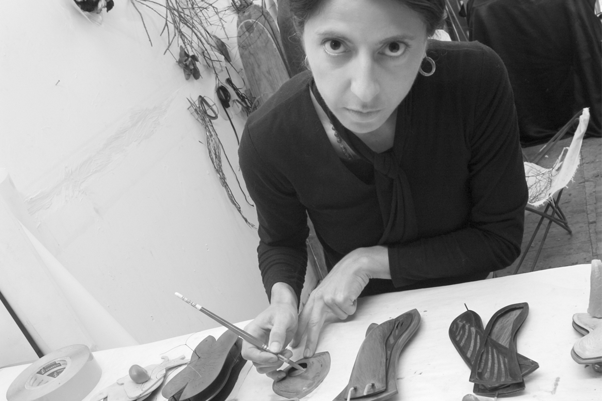 A black and white photograph of a woman at a work bench with a small tool in her hand. She is wearing a dark jumper and is looking intensely at the camera.
