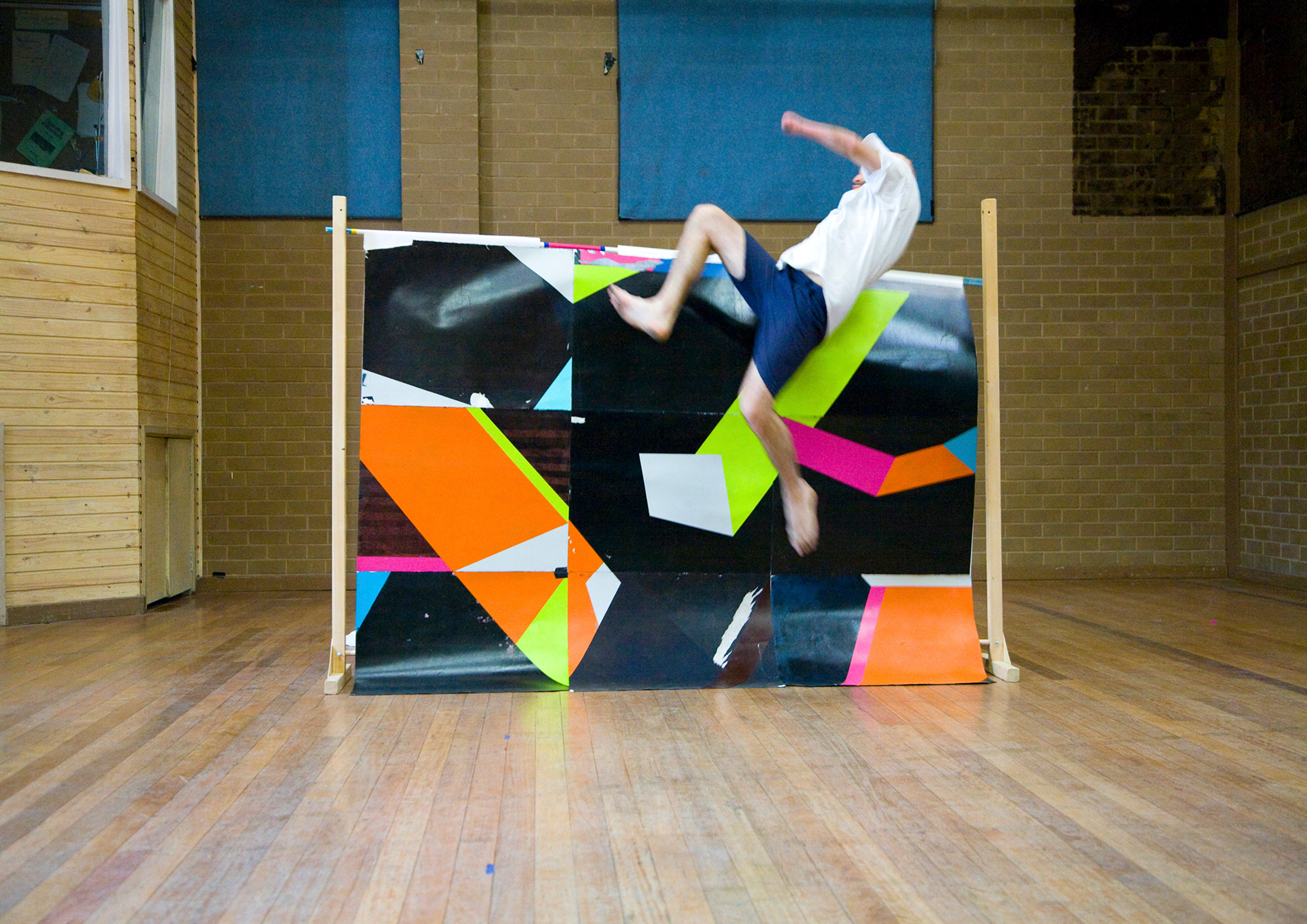A photograph of a man jumping over a screen with a bright abstract pattern in a school gymnasium,