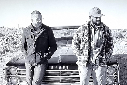 A black and white potgraph of two men leaning against the bonnet of a vintage car.