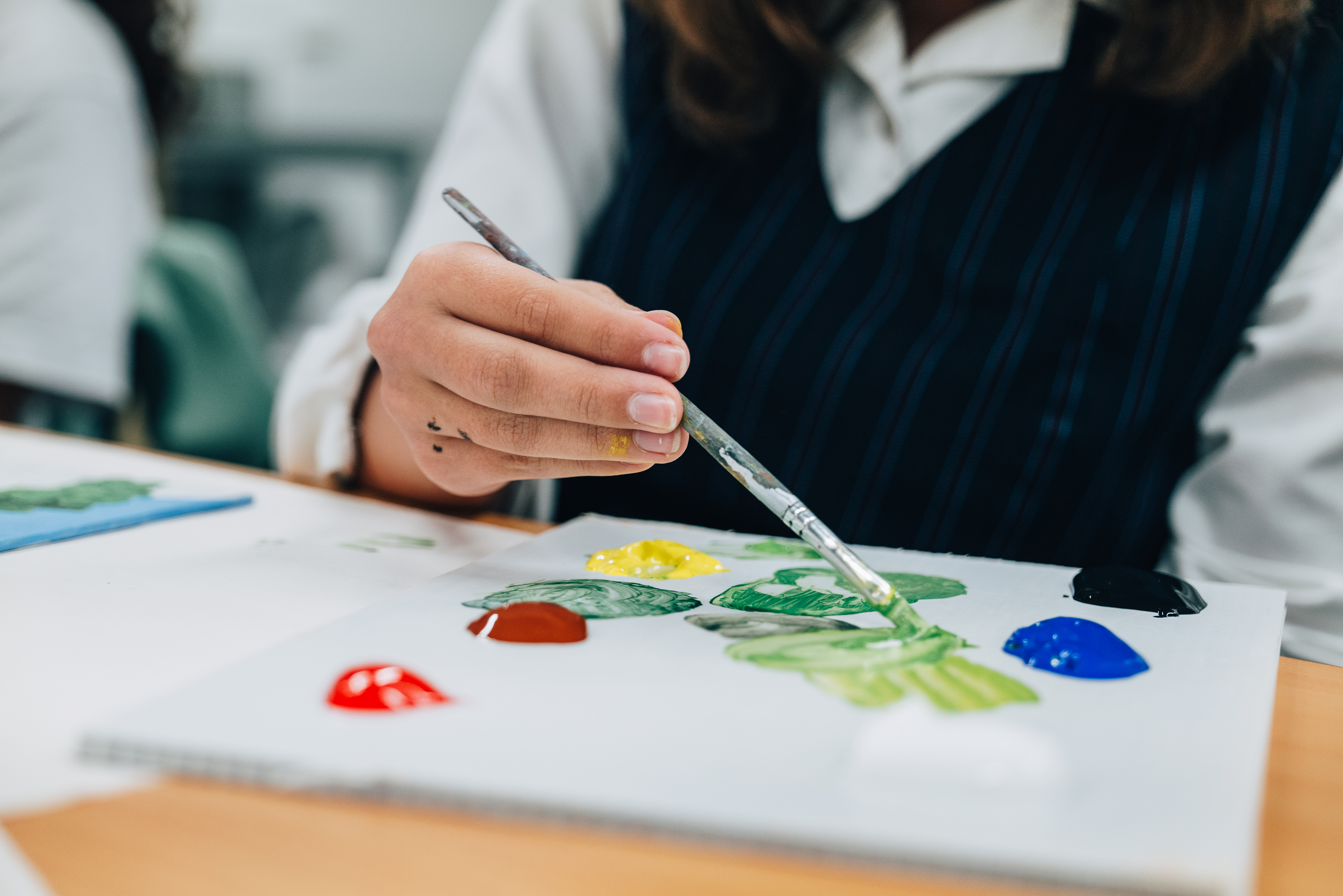 Female student wearing green tartan pinafore and white long sleeve shirt holing paintbrush over palette with large globs of red, yellow and blue paint. She is making the colour green.