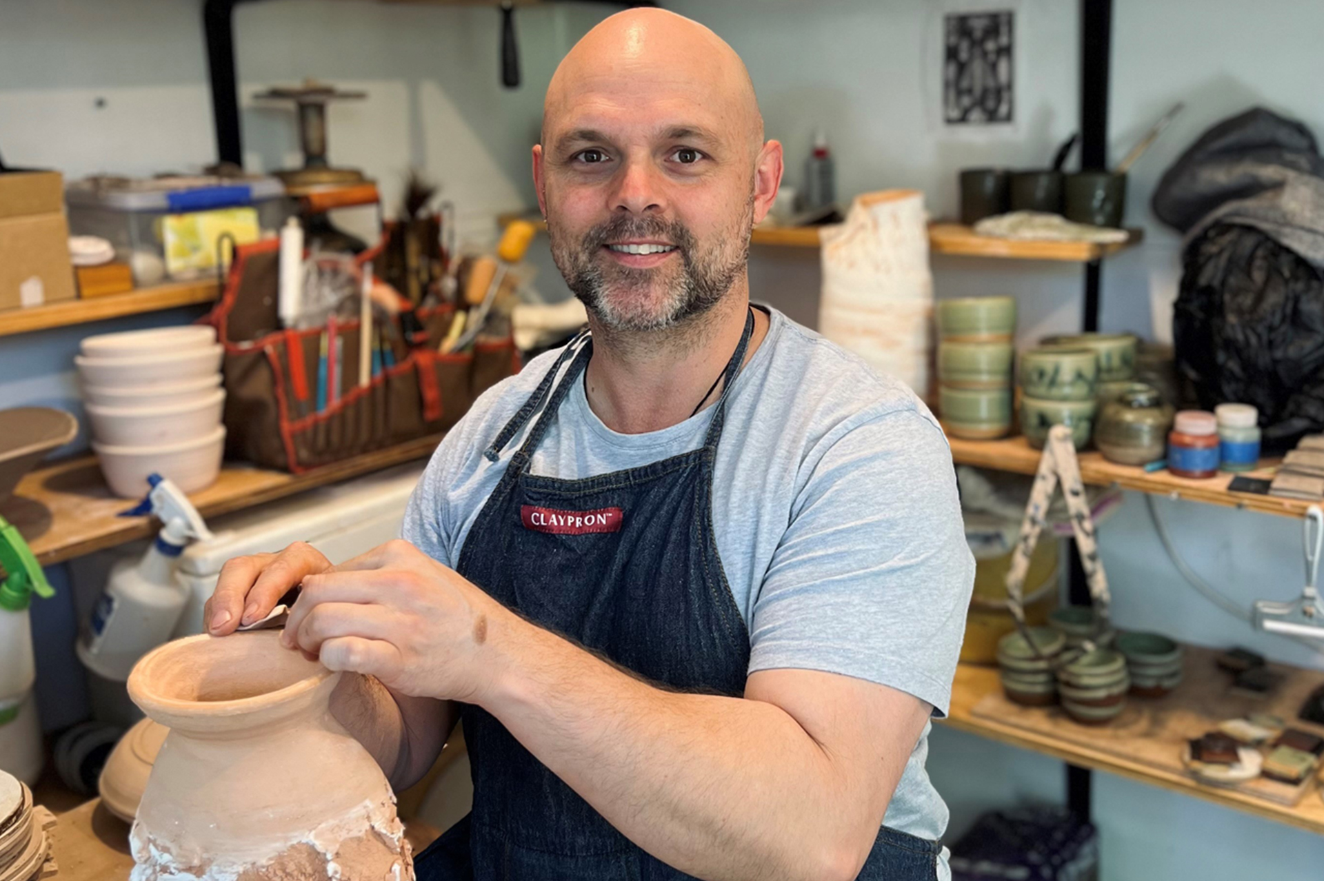 A man wearing a light blue t-shirt and a dark apron sitting in a ceramics studio with his hands resting on a large pot. Ha has no hair and is smiling at the camera.