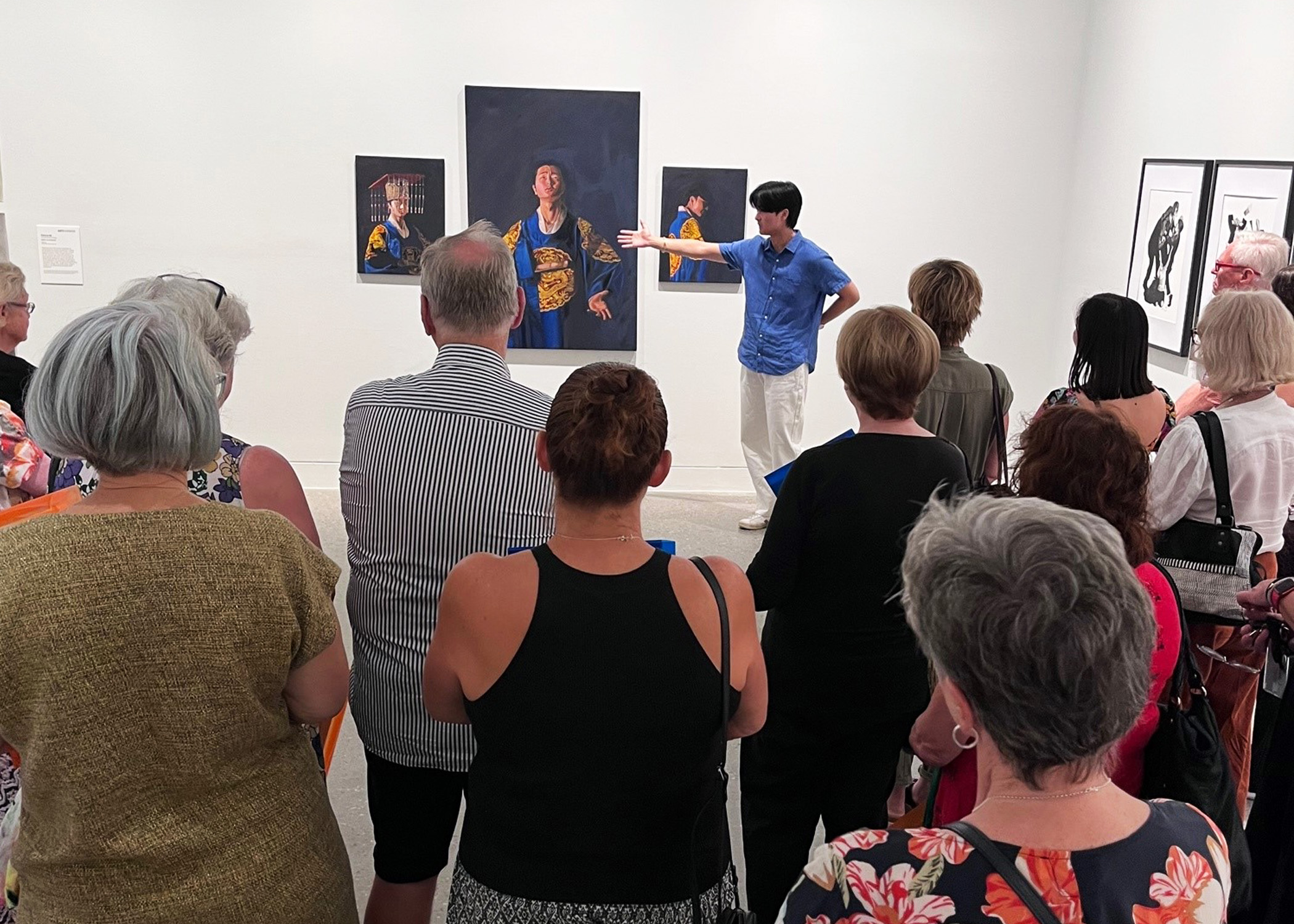 A male HSC student standing in front of a crowd speaking about his painting that is on the wall behind him.