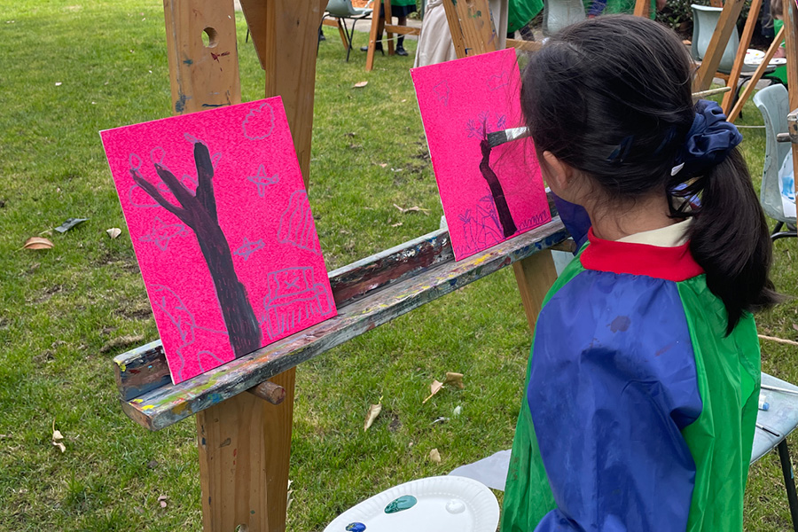 Young female student with dark hair wearing a blue and green paint smock painting a tree on a small hot pink canvas in the gardens. The canvas is on an easel.