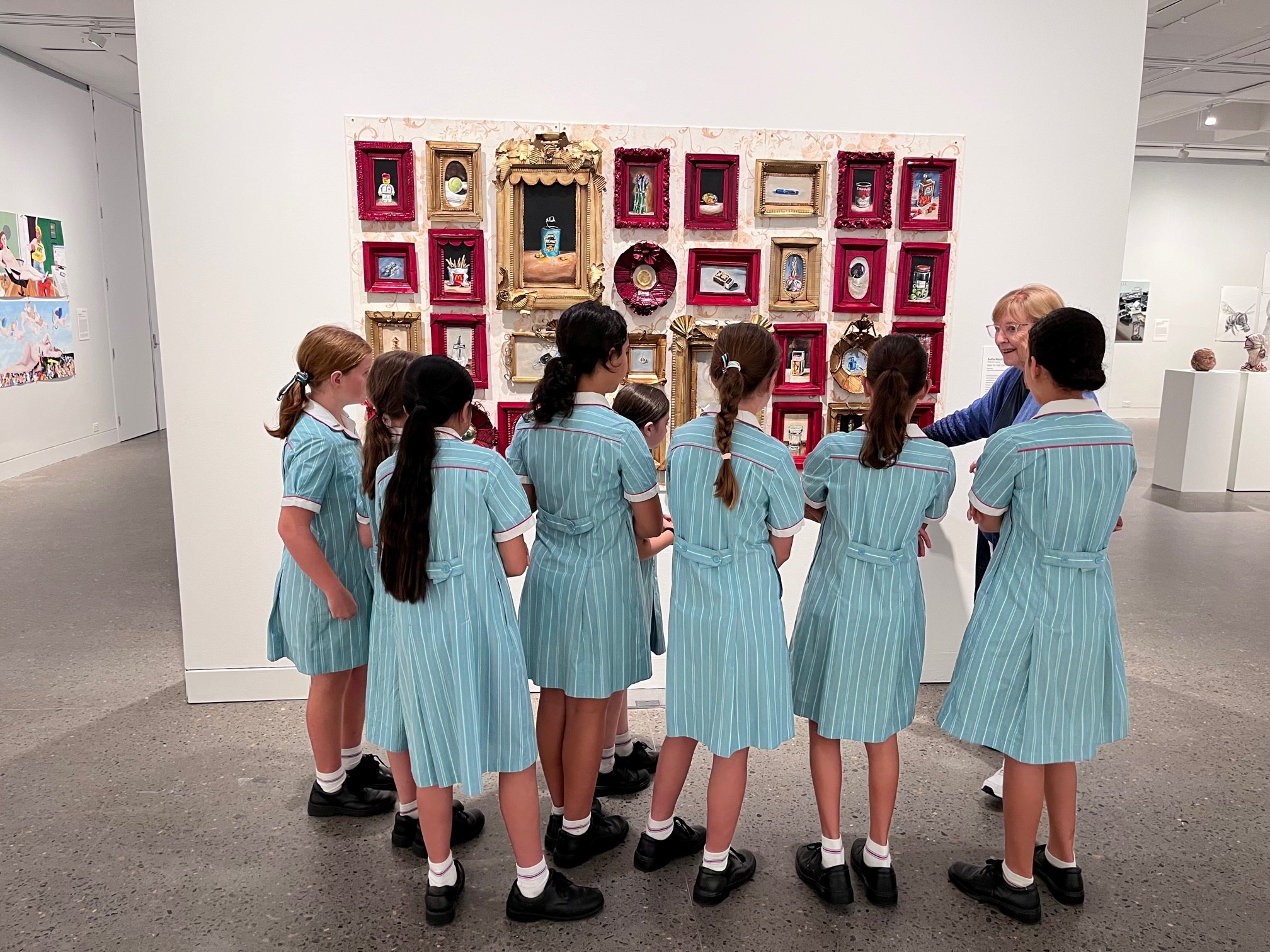 Six female high school students standing in front of an artwork made up of many tiny dark red and gold frames listening to a guide.