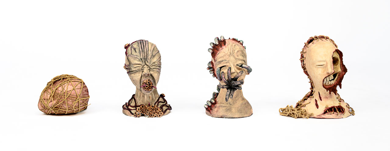 Four grotesque humanoid ceramic heads, slightly monstrous, all displaying physical trauma and wounds.