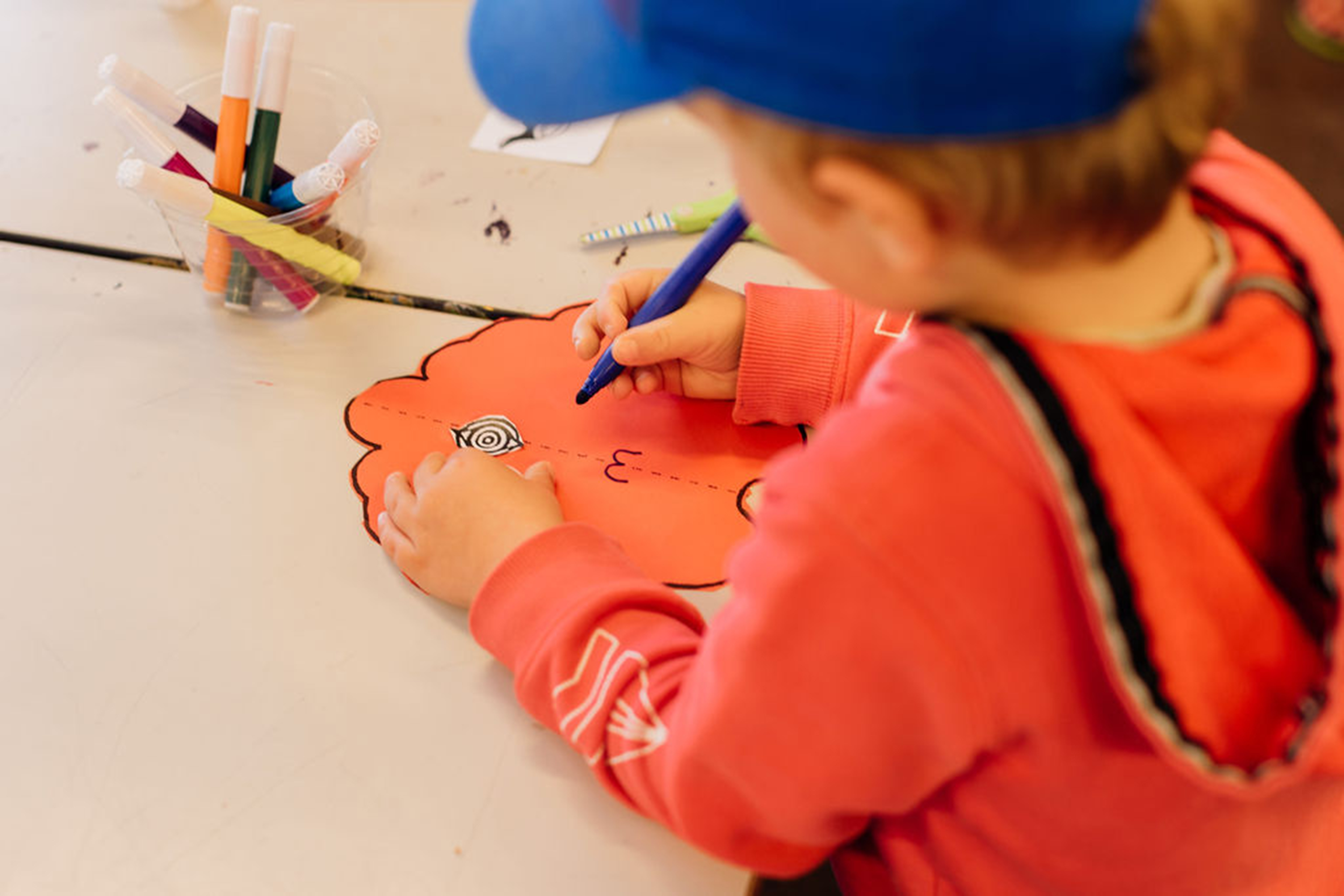 Young boy in orange jumper and blue hat colouring in.