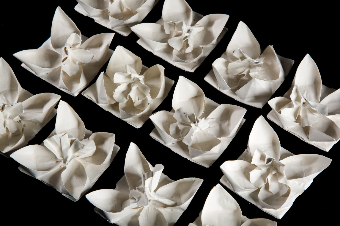 12 small wite ceramic works that are so delicate the look like origami lotus flowers. They are arranged in three rows of four and are on a black background.