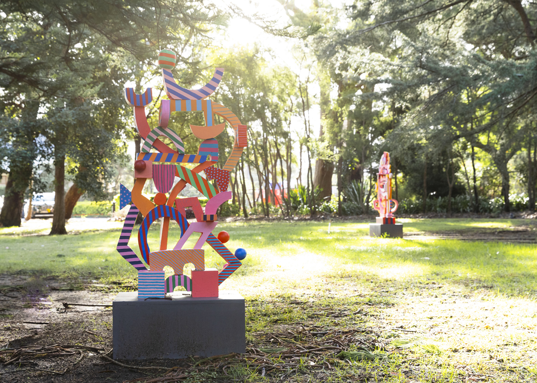 Three large colourful sculptures featuring different lines, shapes and patterns sitting among trees with the sun shining behind them.