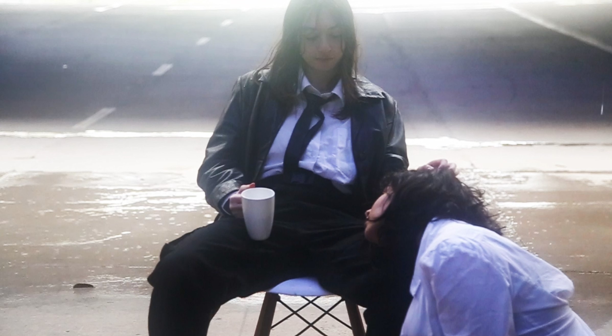 Film still featuring female person wearing suit sitting in a chair with another female person with their head in he lap. 