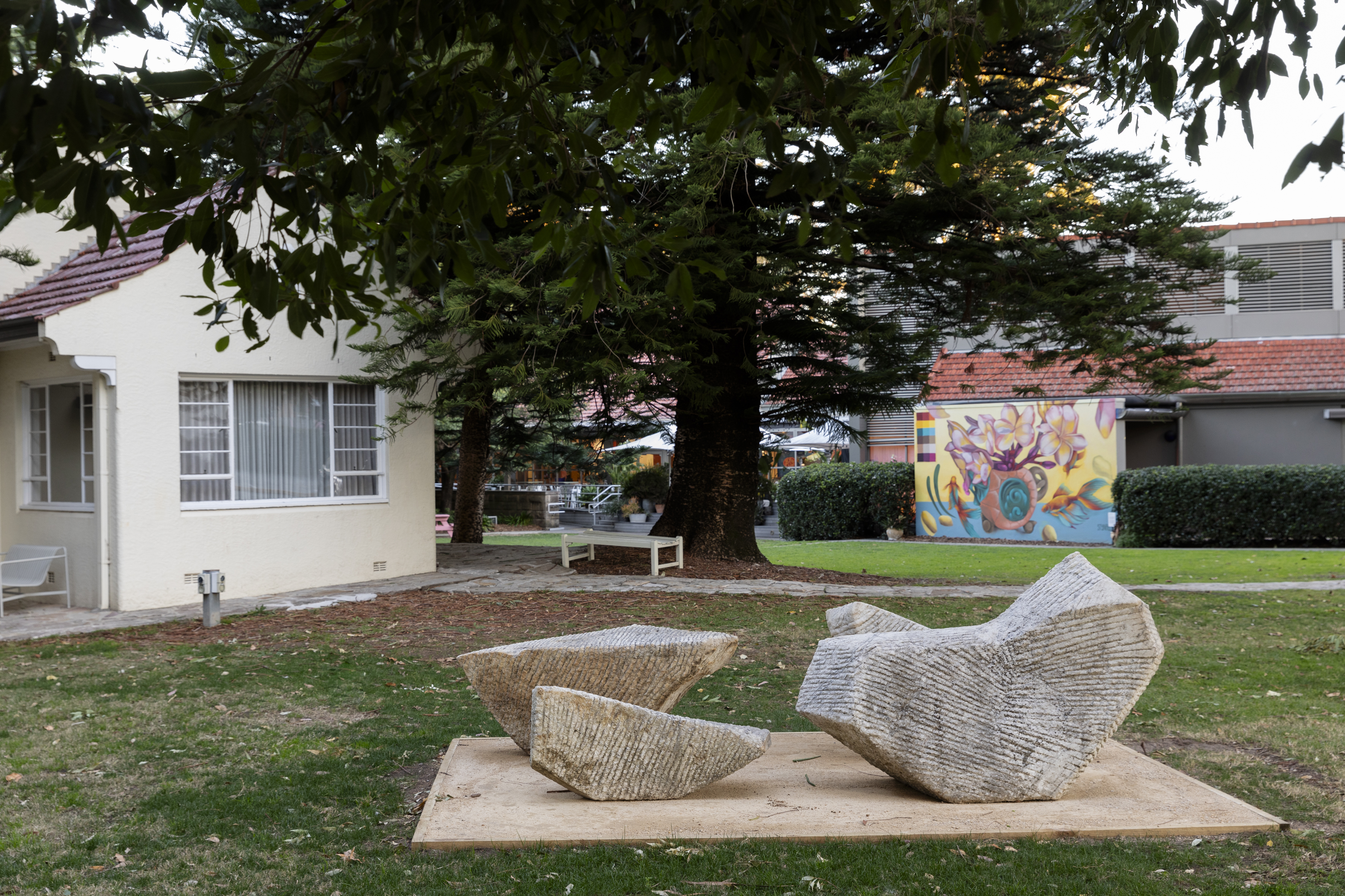 Three large organic stone shapes sitting on a square of earth coloured crushed gravel in front of the Hazelhurst cottage. The Arts Centre and the colourful mural are in the background.