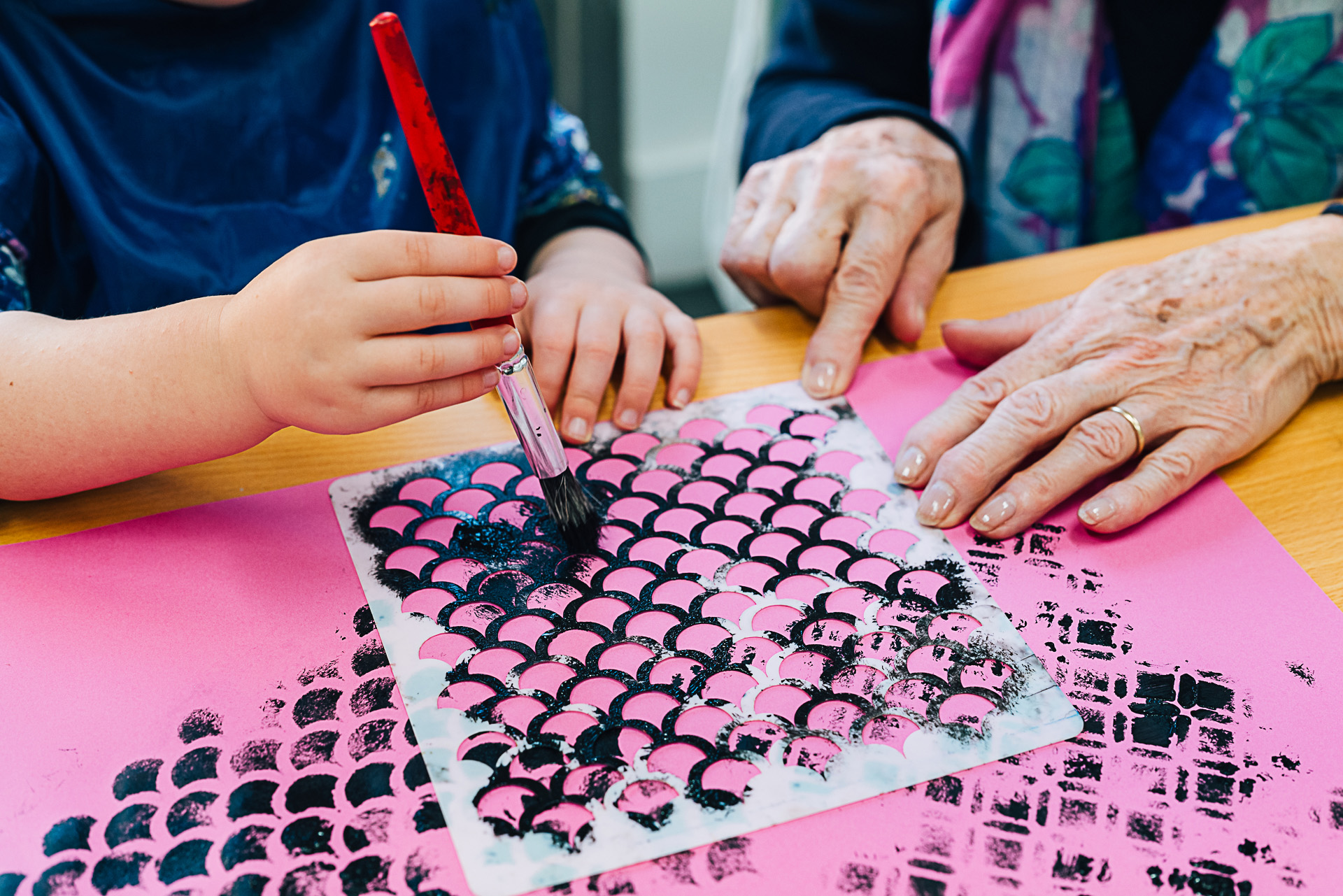 Granddaughter and grandmother painting on pink paper using a stencil and black paint.