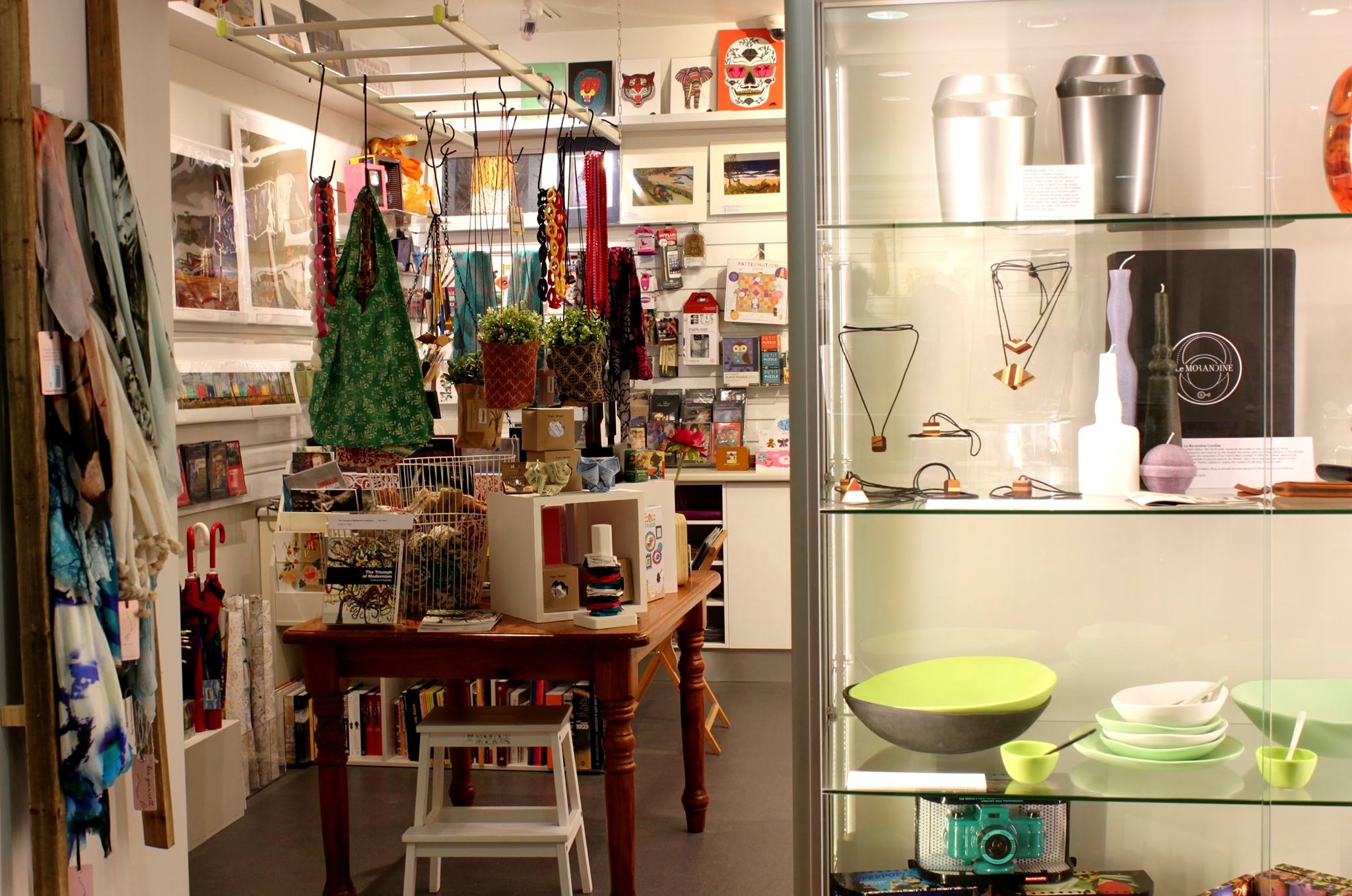 Looking through the doorway of a small white walled shop space, around the walls are artworks and paper goods and their is a talbe of wares in the centre, and a ladder with bags hanging from the roof. On the left of the door is a rack of colourful scarves and on the right is a lit up glass cabinet showcasing jewellery and ceramics.