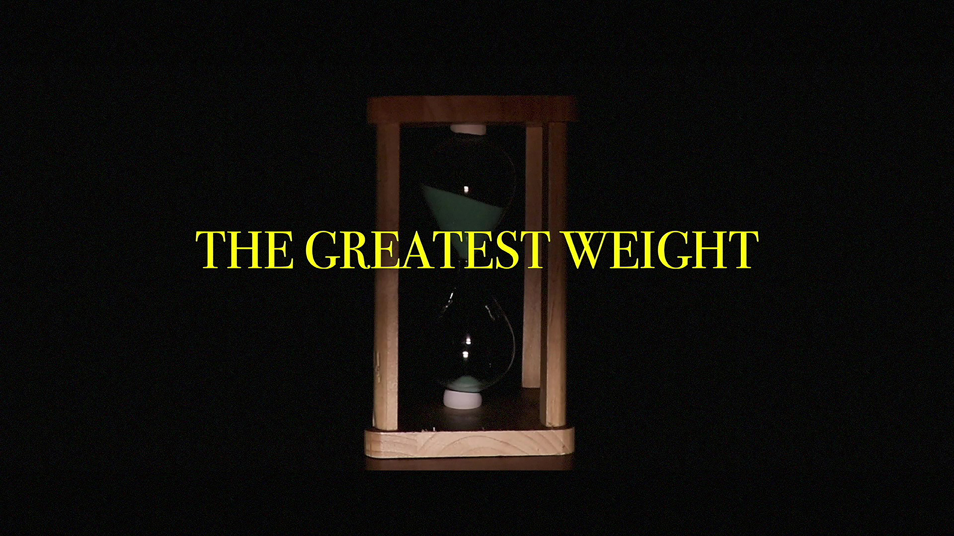 A still from a film showing an hour glass with the title of the work "The Greatest Weight" overlayed in yellow.