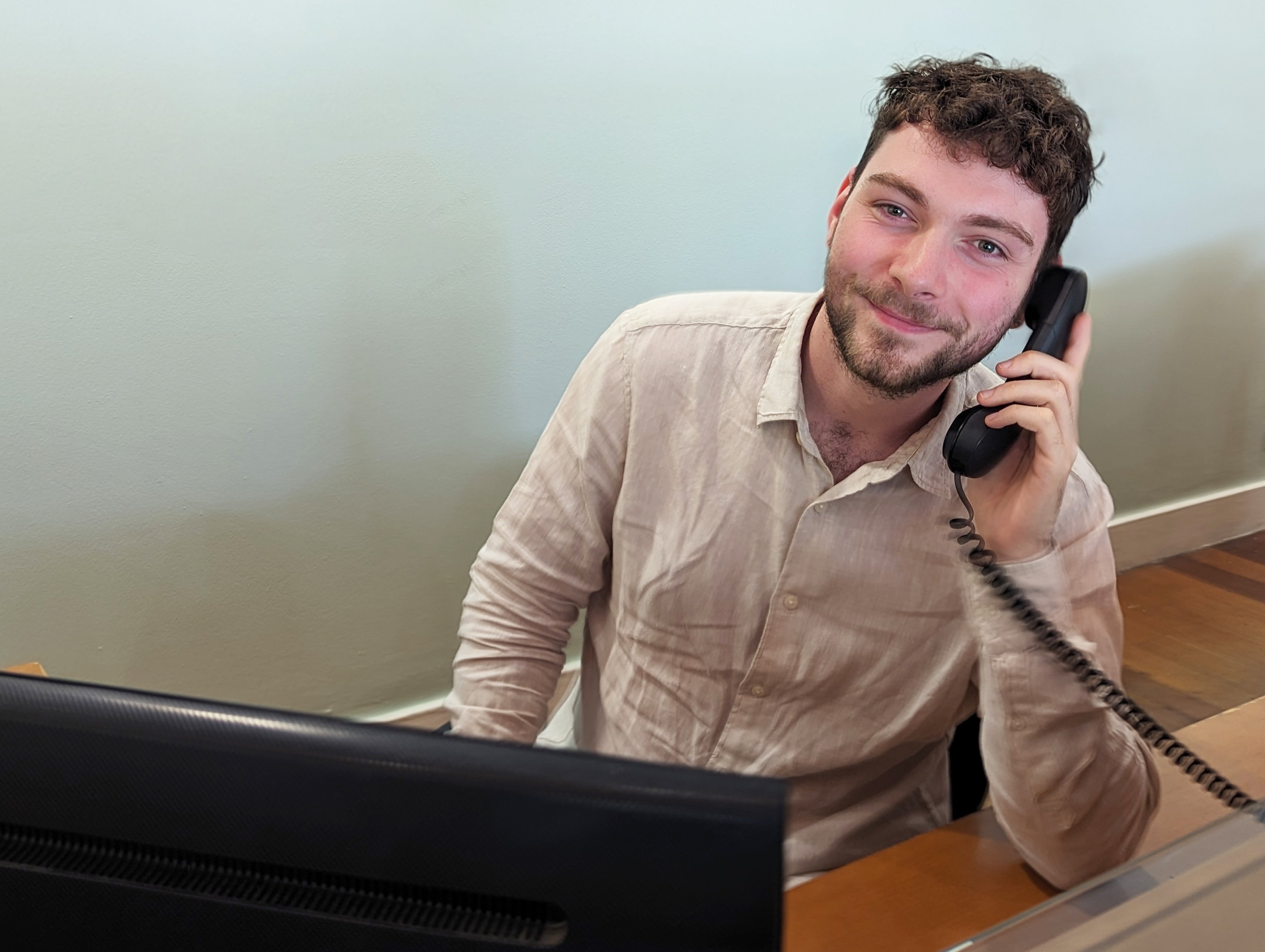 A youn man sitting at a computer, holding a phone, smiling at the camera.