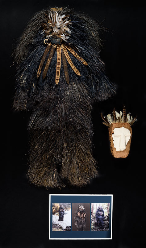 A feathery looking full body costume and elaborate mask in dark, neutral tones.
