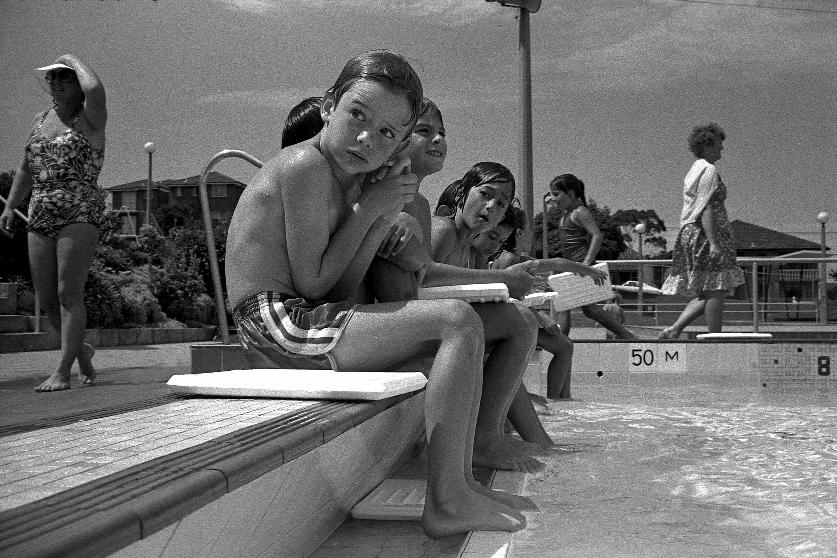 A black and white photograph of children sitting on the edge of a public swimming pool.