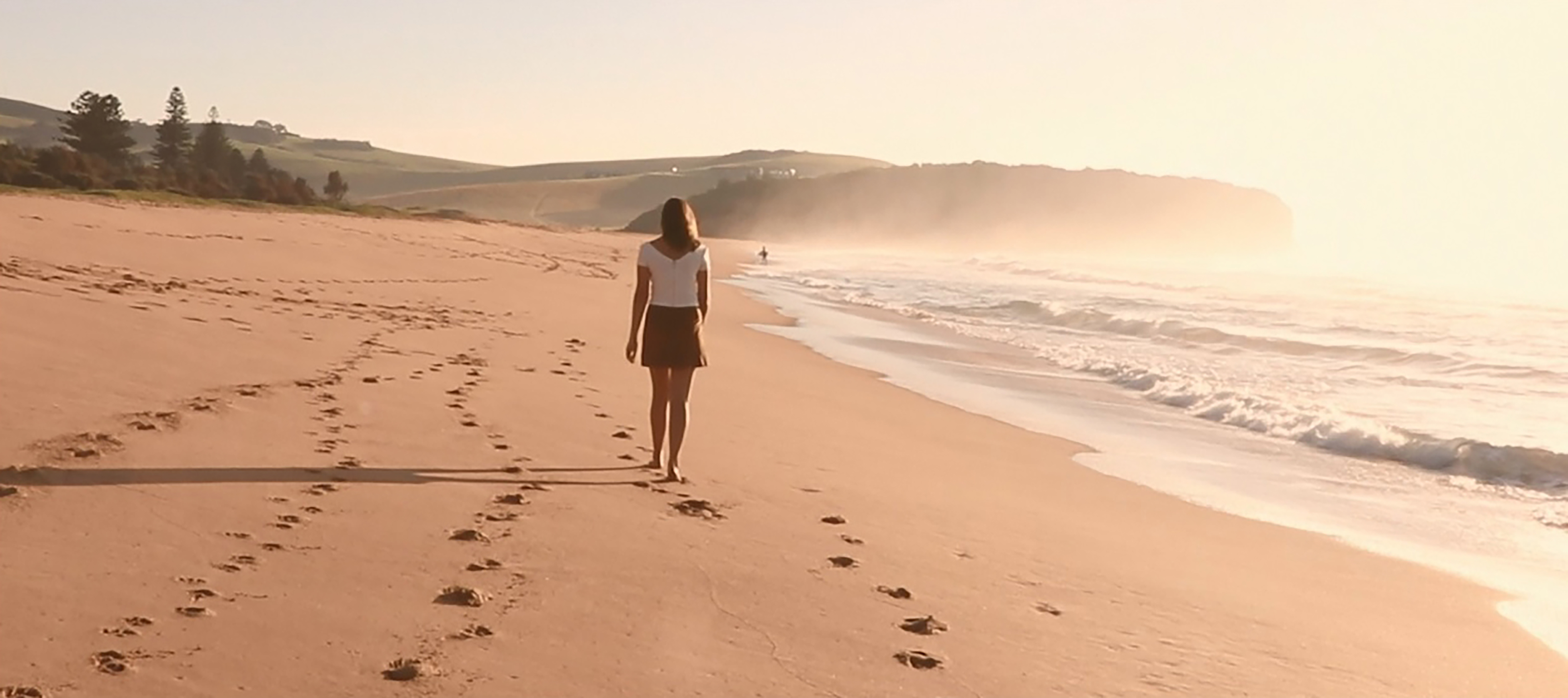 A still image of a female person walking along a beach at sunset.