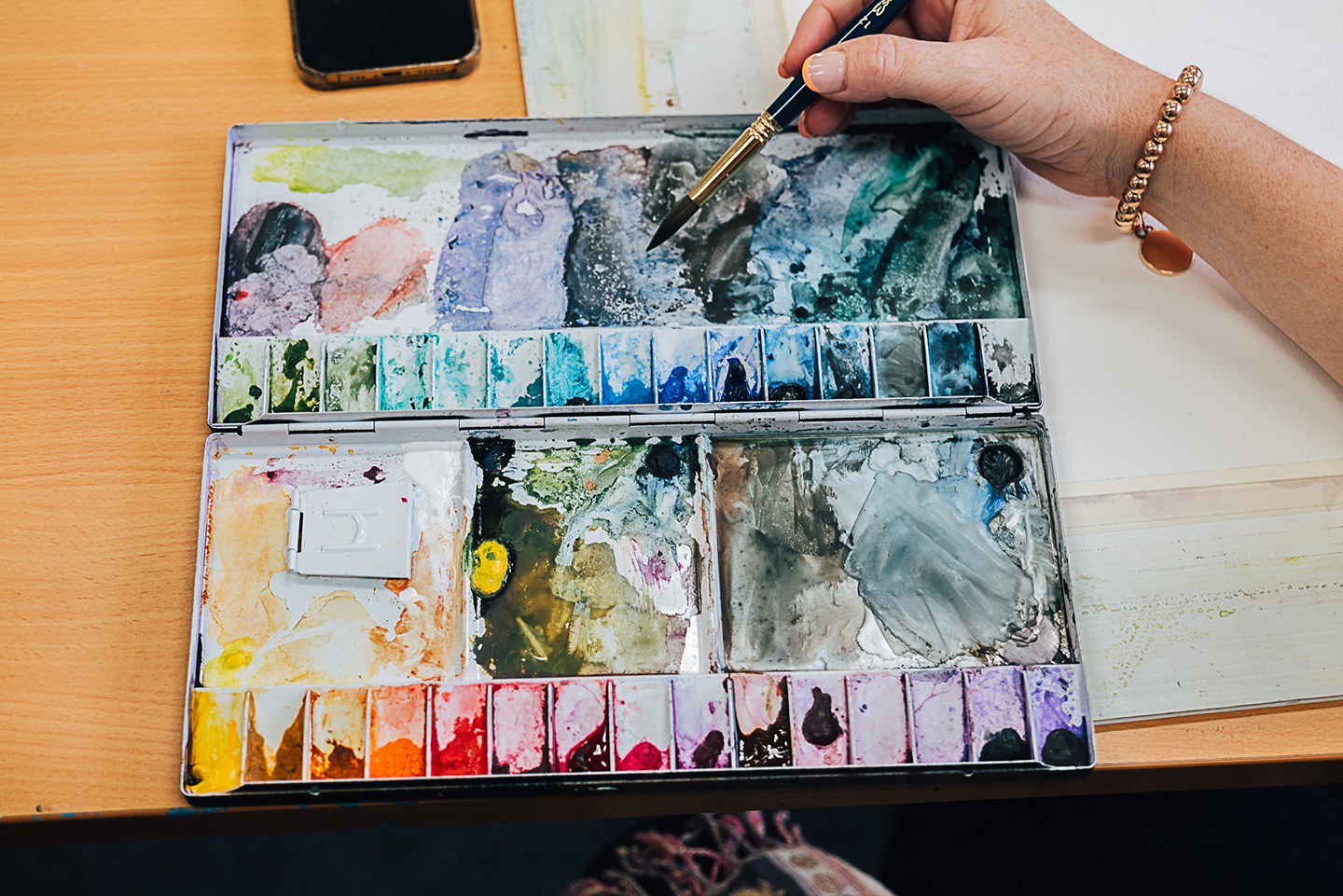 A ladies hand holding a paintbrush over a watercolour palette.