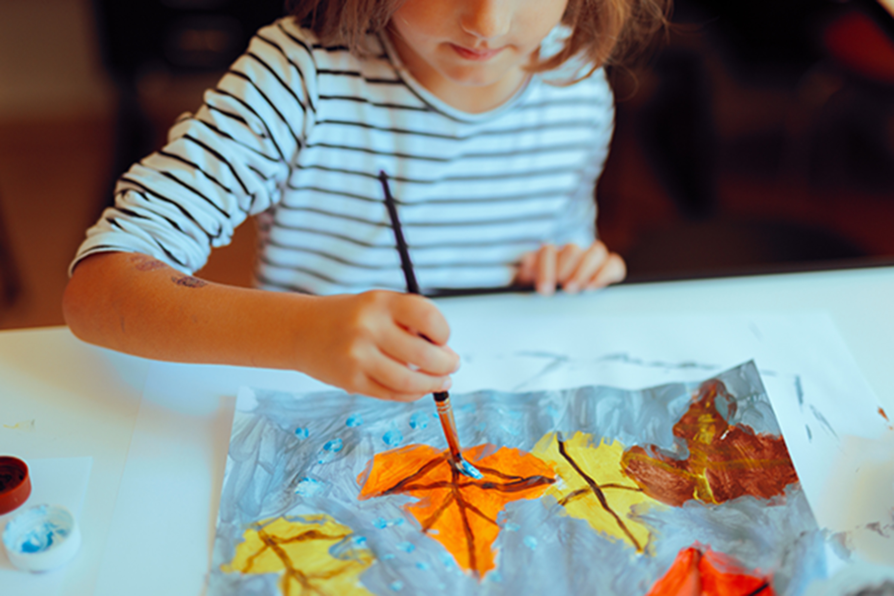 A school aged girl in a black and white striped top painting autumn leaves on a piece of paper.