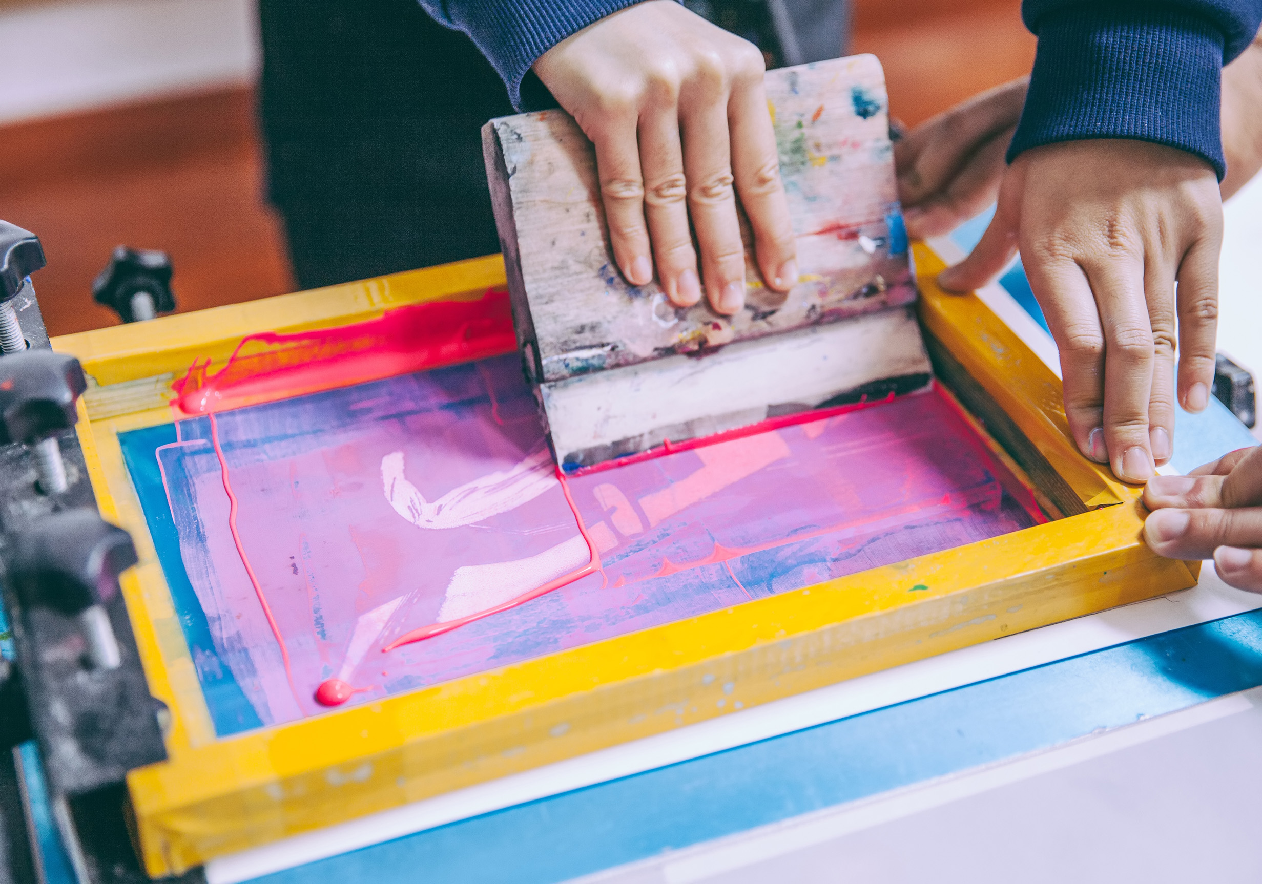 Two people's hands using screen printing equipment with hot pink ink.