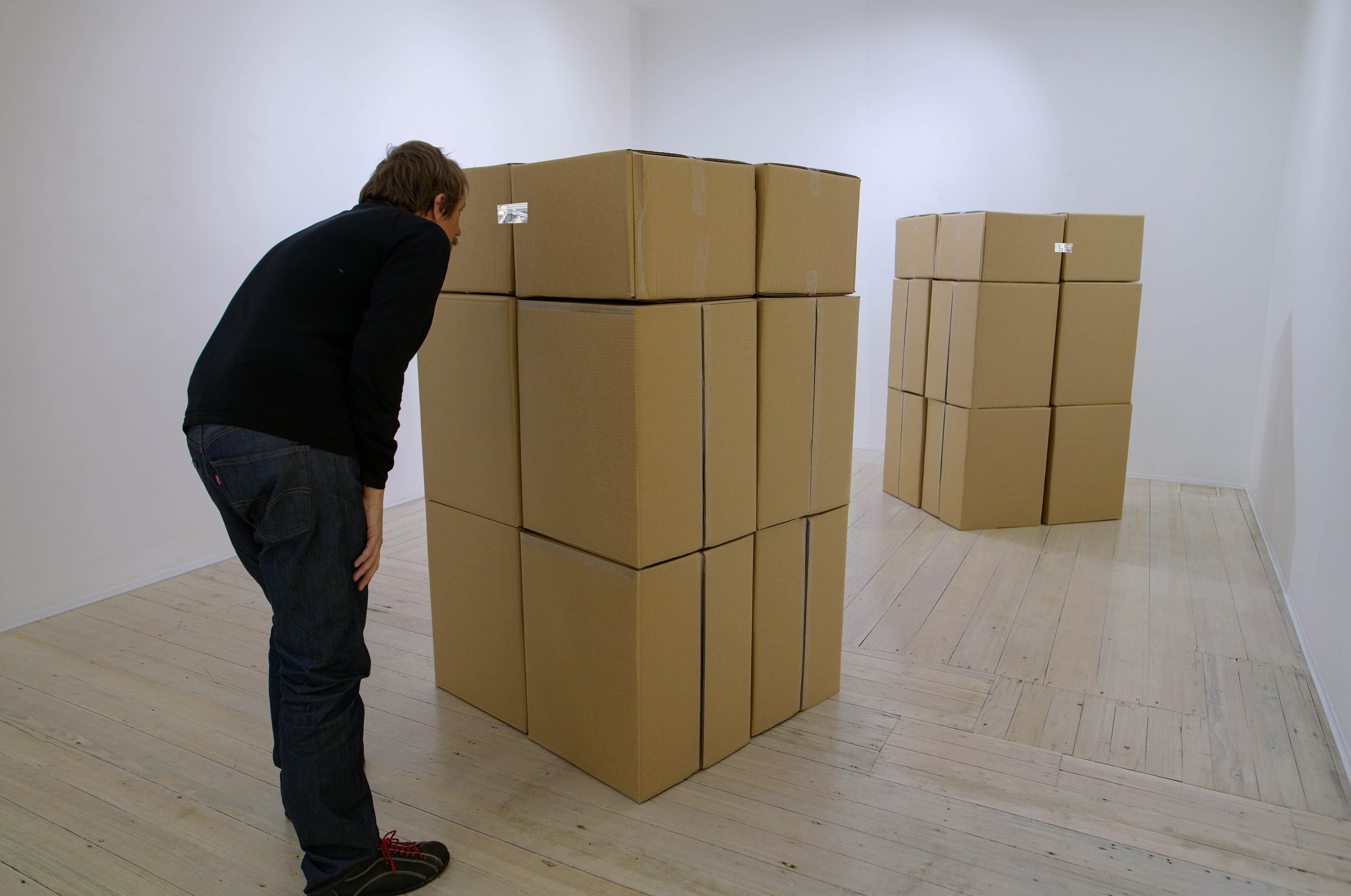 Two large stacks of plain brown cardboard boxes in a gallery space. There is a man dressed in dark clothing bending slightly to look into a small window at the top of the stack closest to the camera.